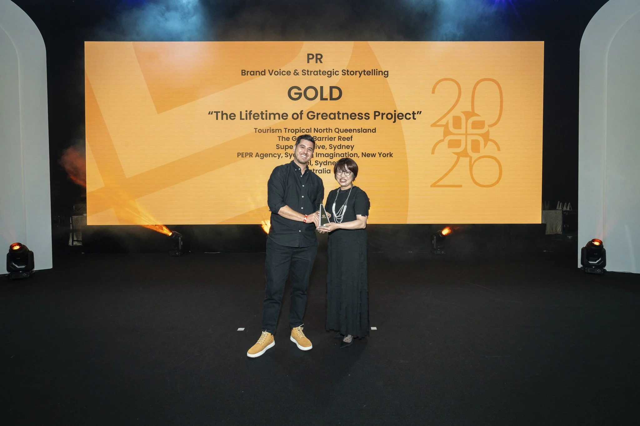 Two people standing on a stage in front of a large screen, holding an award. The screen displays information about an award for PR, Brand Voice & Strategic Storytelling, Gold category, titled 'The Lifetime of Greatness Project.' The background is dark with stage lights.