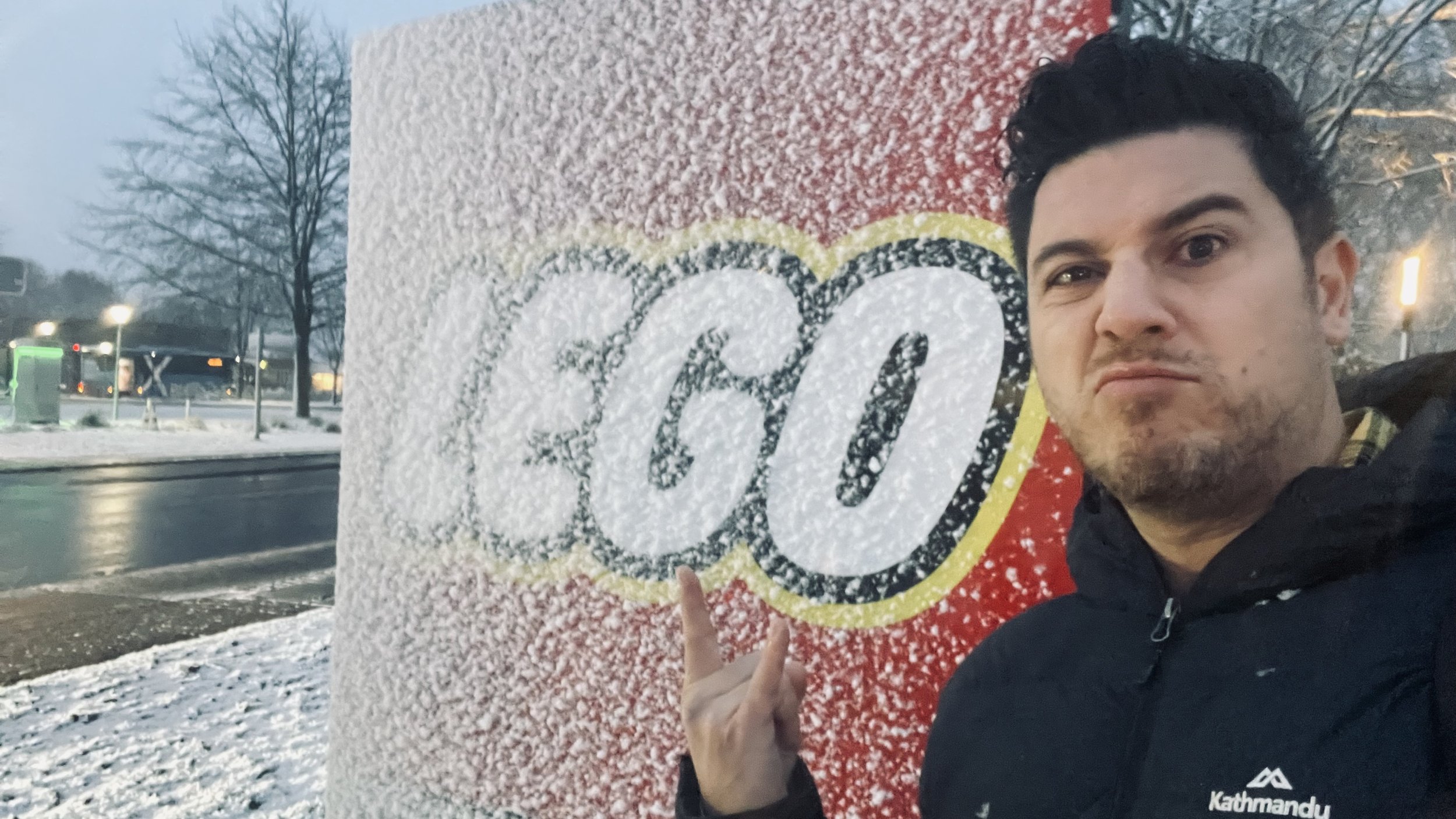 A man with dark hair and a beard taking a selfie outdoors in snowy weather, standing next to a large colorful sign with snow on it, featuring the logo of the snack brand Doritos, with trees and streetlights in the background.