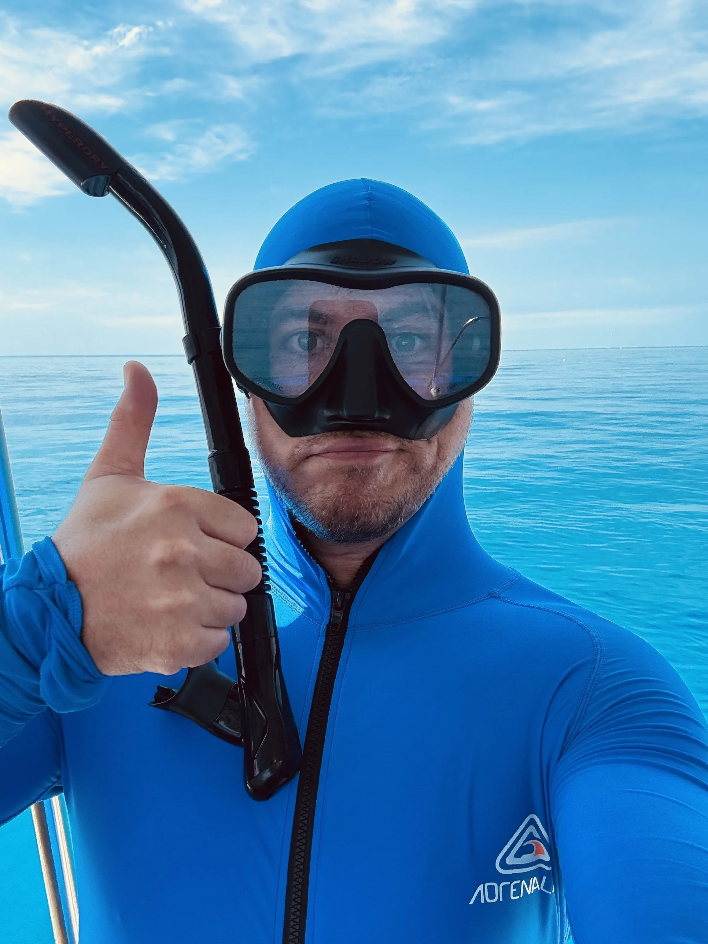 A man in a blue hooded jacket and a blue cap, wearing snorkeling gear with a mask and snorkel, takes a selfie with an ocean in the background, and is giving a thumbs-up.