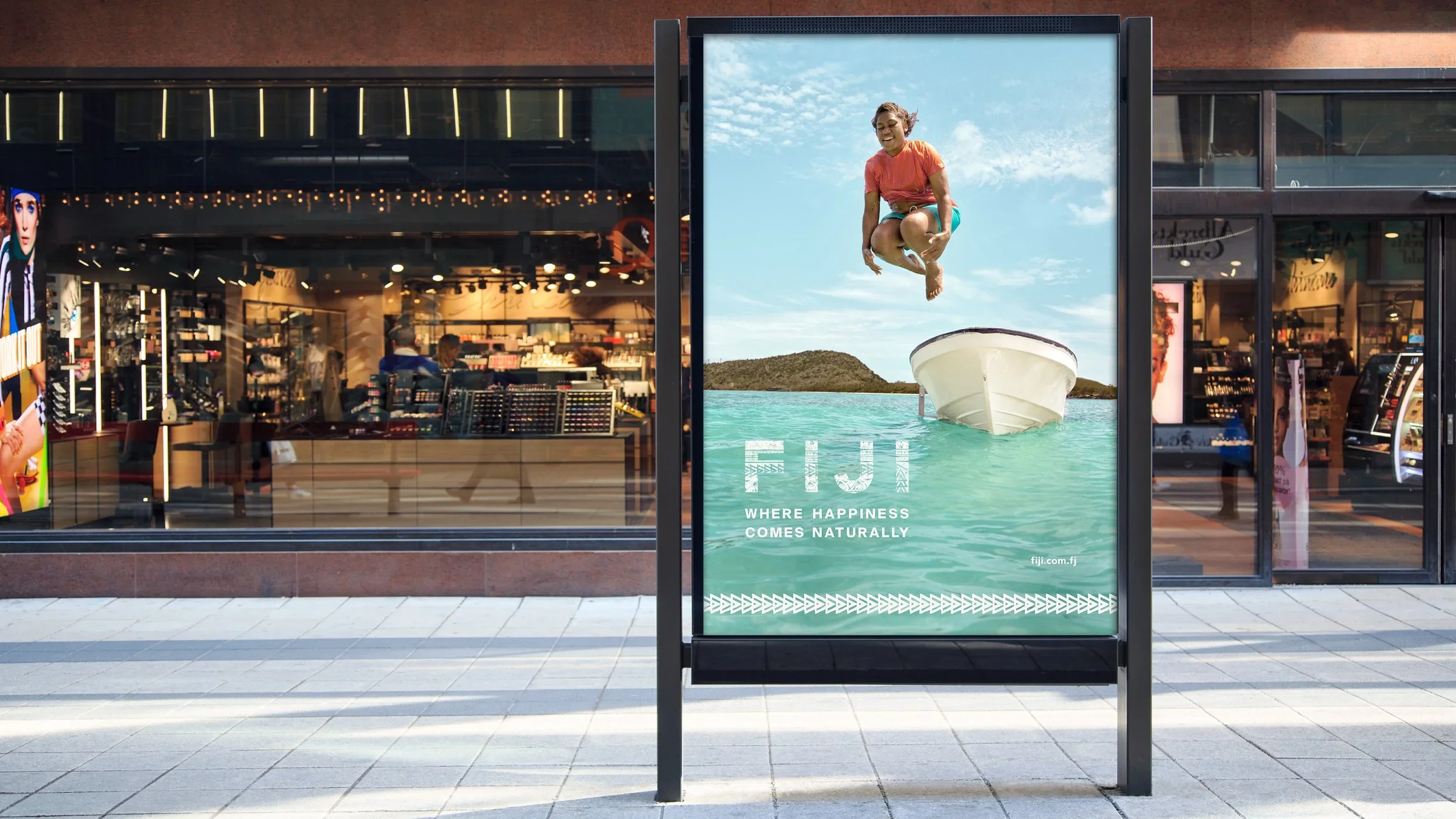 Advertising poster on a sidewalk showing a woman jumping over water with a boat and blue sky background, with the text 'WHERE HAPPINESS COMES NATURALLY'.