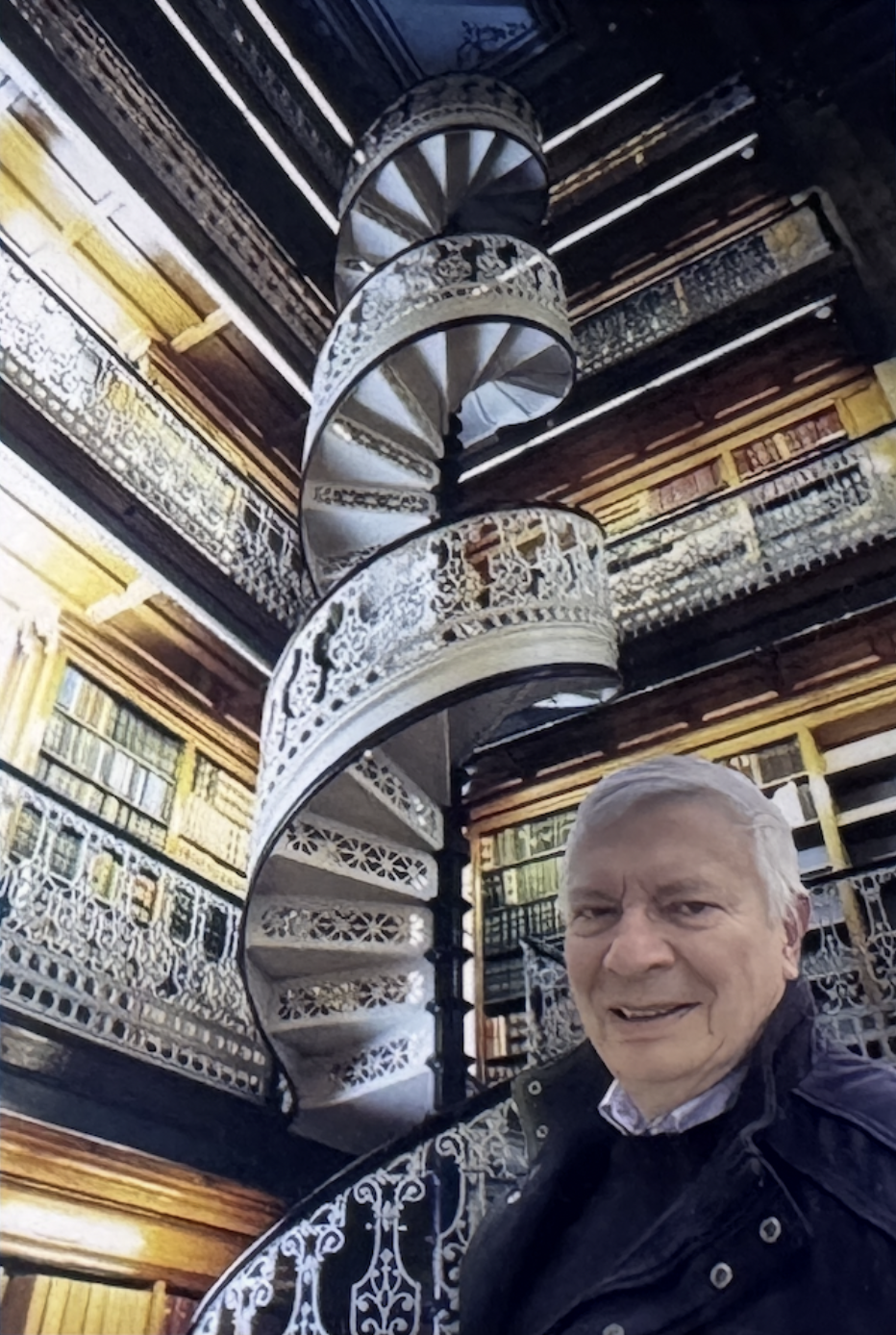 Un homme avec des cheveux gris portant une veste noire, debout devant une bibliothèque ancienne et une escalier en colimaçon en fer forgé blanc avec des motifs décoratifs.