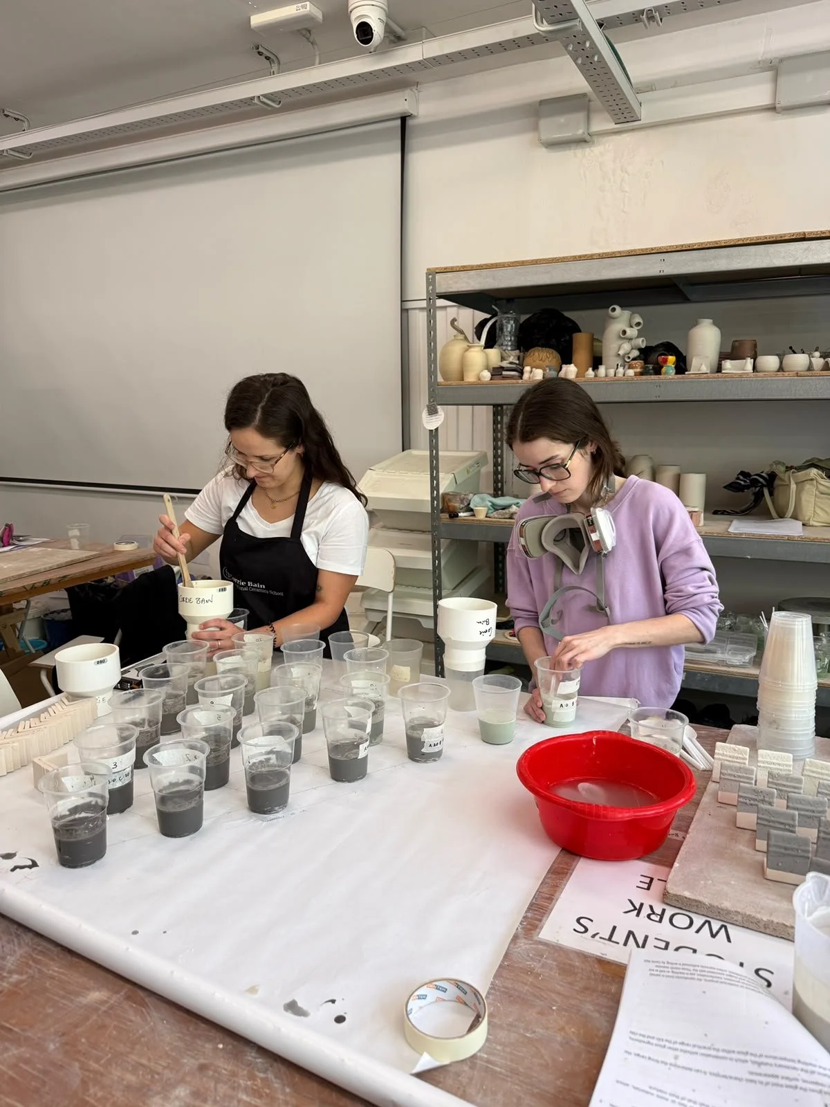 Two women working on ceramic painting or glazing, with numerous cups of glaze or paint on the table, in a ceramics studio with shelves of ceramic pieces and supplies in the background.