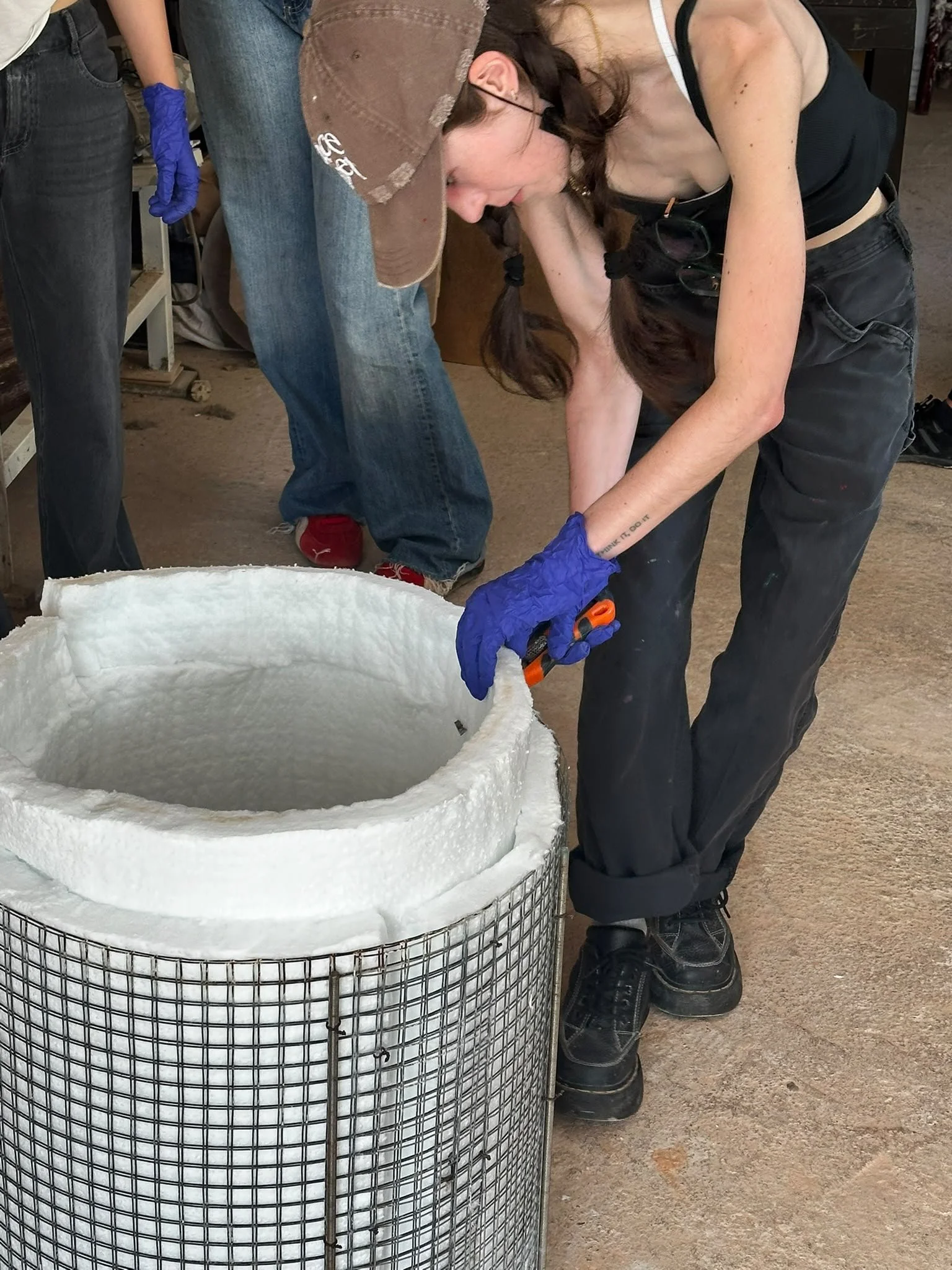A woman with long brown hair, wearing a brown baseball cap, black tank top, and black pants, is bent over working on a project with a large circular well or container. She is wearing blue gloves and black boots. A person standing nearby is wearing je