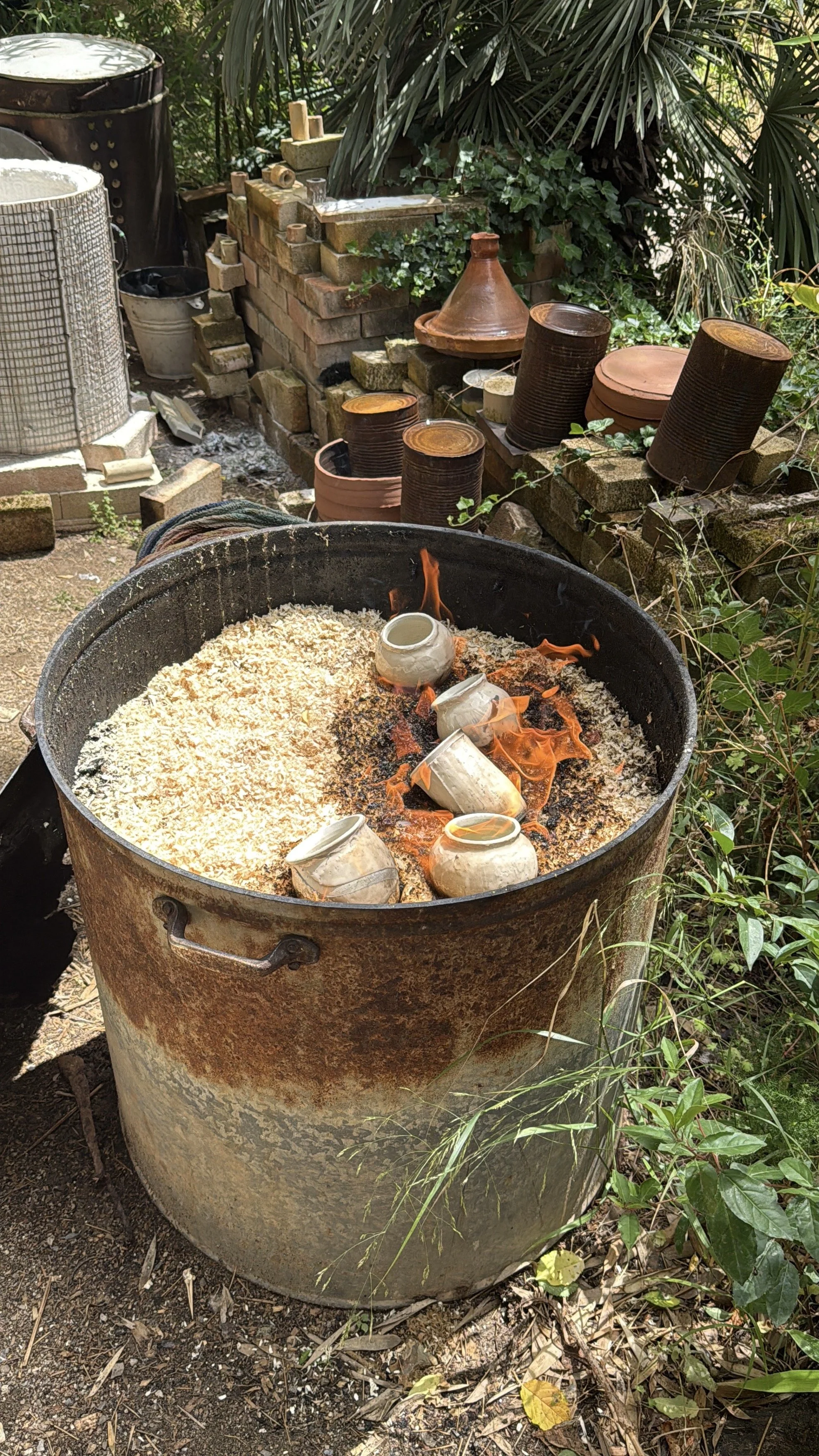 Outdoor scene with a large rusted metal barrel filled with shredded wood or straw, burning with a small flame, and ceramic cups placed on top of the fire. In the background, there is a brick wall with various clay pots and gardening items, surrounded