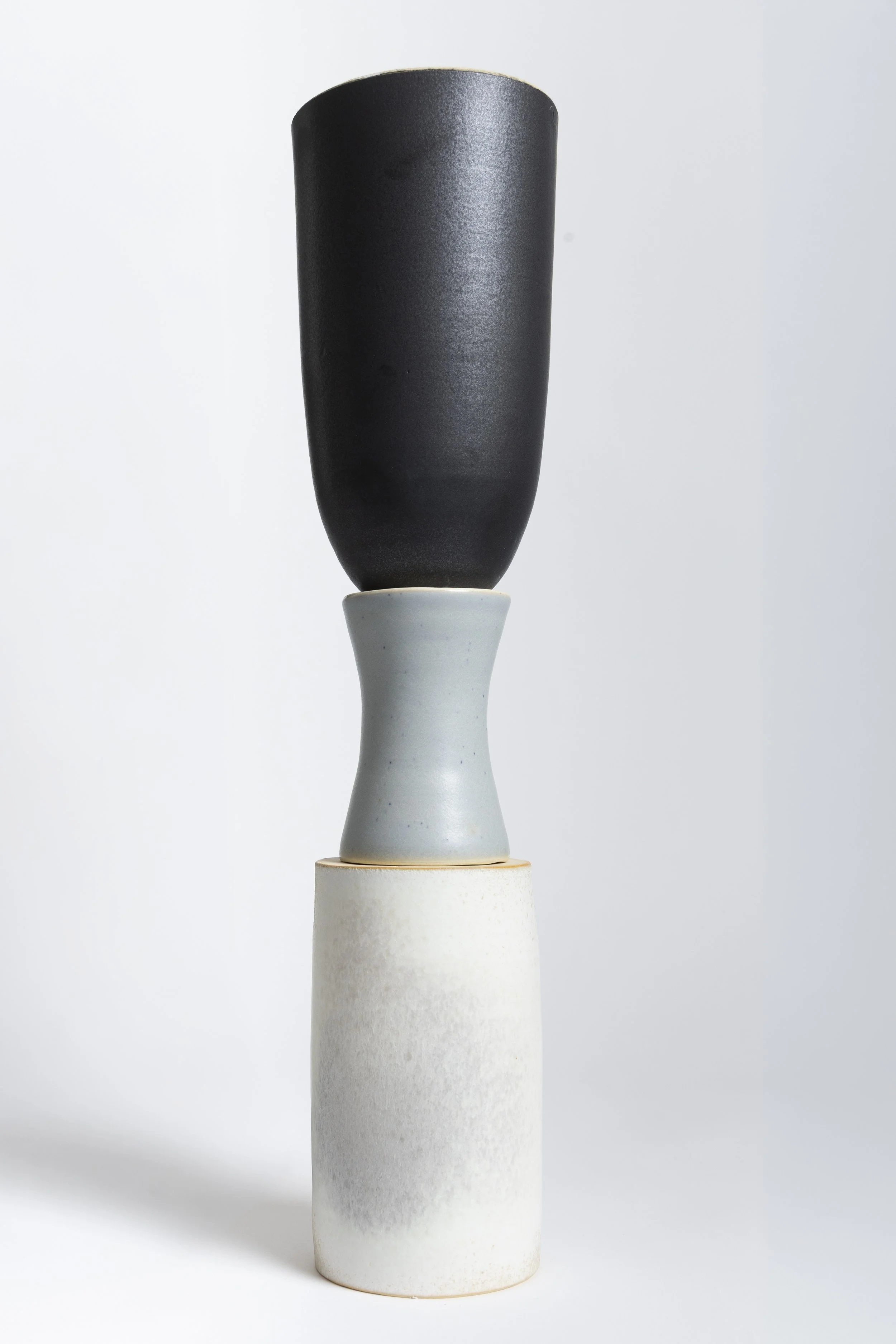 Three stacked ceramic vases, with the top vase being black, the middle vase light gray, and the bottom vase off-white, on a white background.