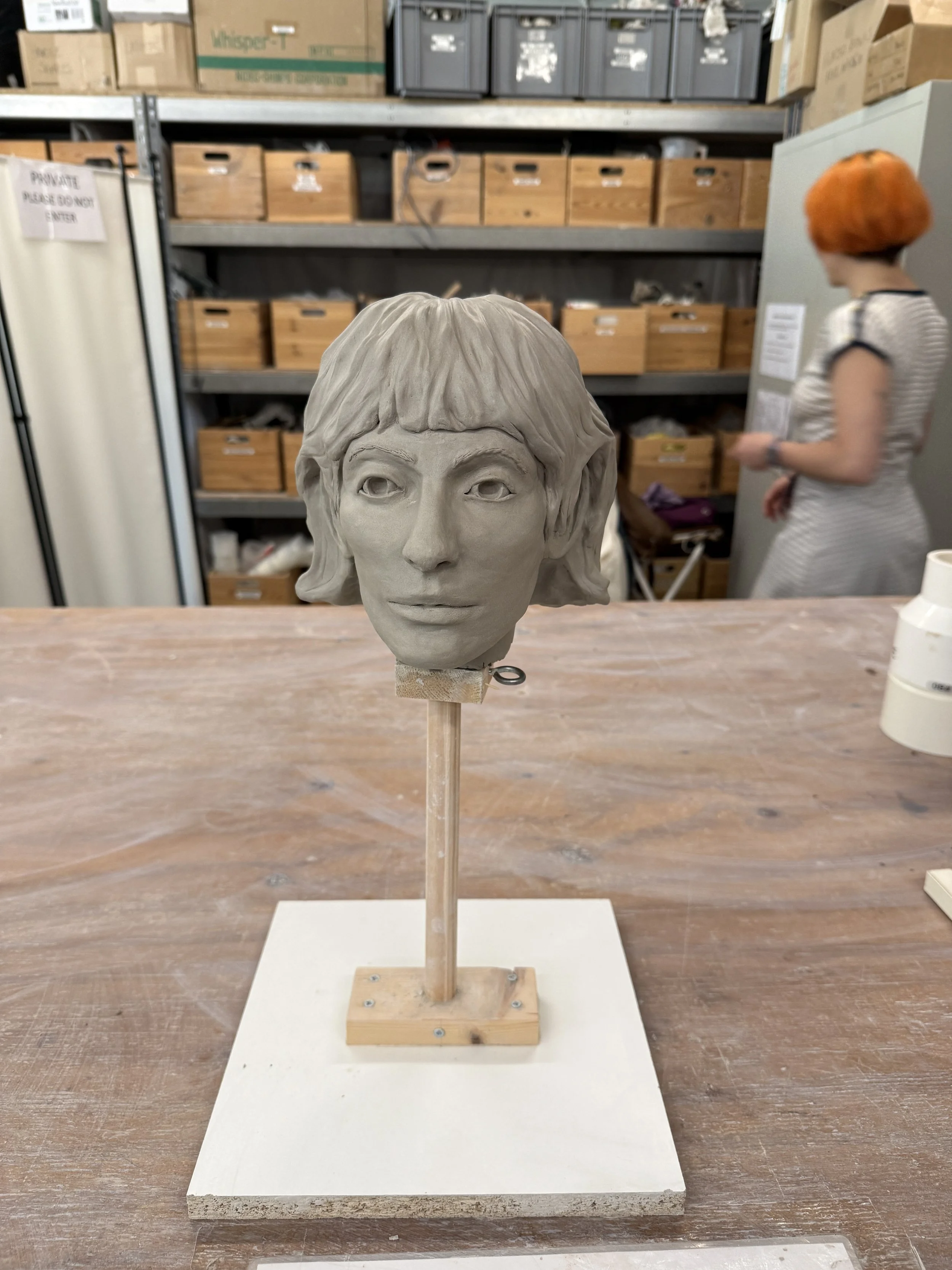 A clay bust sculpture of a woman with short hair on a wooden stand, placed on a table in a workshop or storage room.