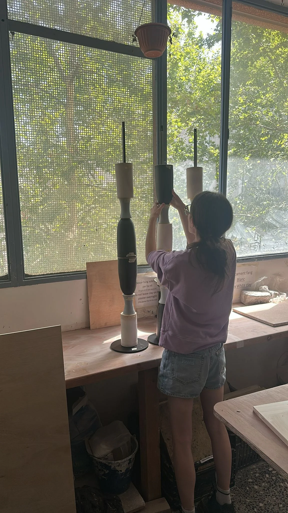 A girl with black hair, wearing glasses, a purple hoodie, and denim shorts, is arranging items on a tiered display of candles made from white, black, and gray materials. She is standing at a wooden countertop near a window with metal grid and greener