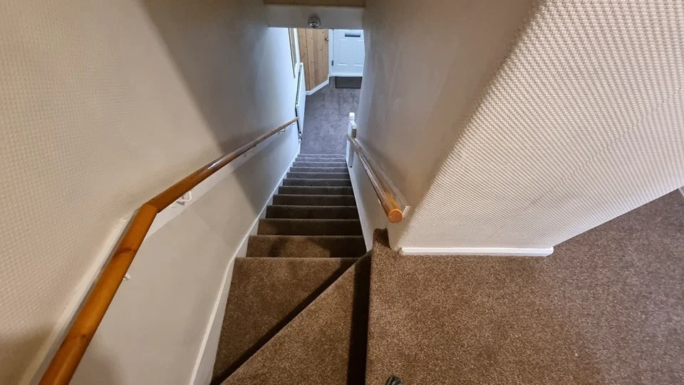 View looking down a carpeted staircase with wooden handrails on both sides, leading to a landing and a door at the bottom.