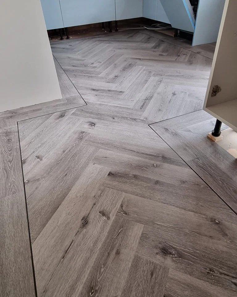 Close-up of a wooden herringbone pattern floor in a modern room with light gray cabinets and white walls.