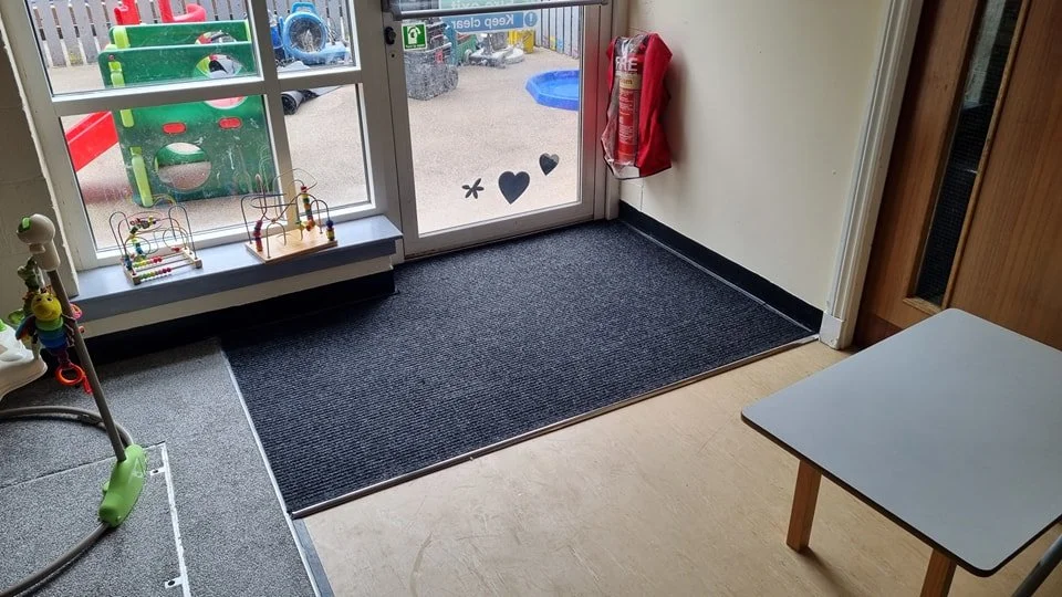 Indoor area with a glass door leading outside, with playground equipment like a slide and sandbox visible. There are toys and a small table with supplies near the door.