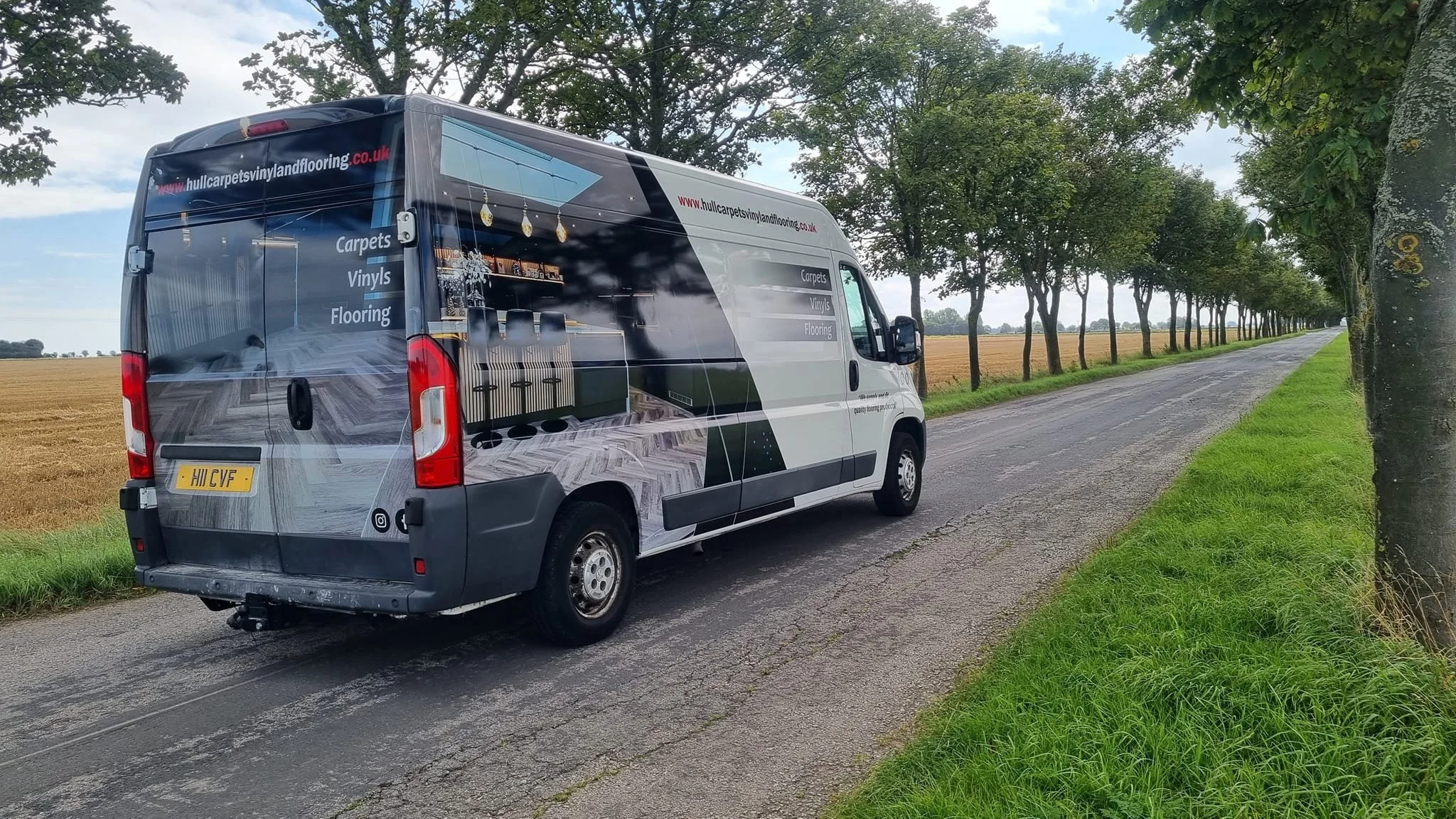 A vinyl-covered delivery van parked alongside a rural road with trees and a field in the background.