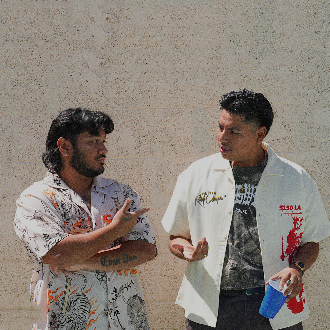 Two men engaging in conversation against a beige brick wall, one with long dark hair and tattoos, wearing a patterned shirt, and the other with short dark hair, wearing a white jacket with red detailing, holding a blue cup. REALIFEKING