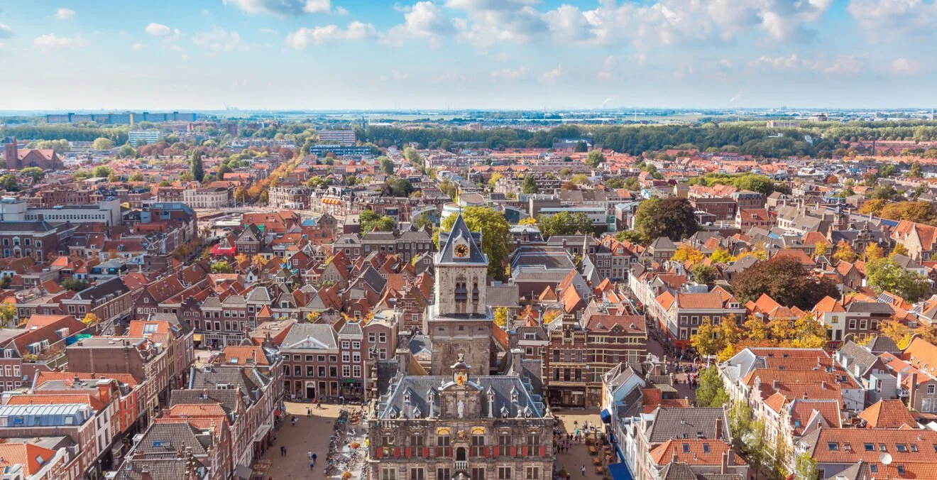 Uitzicht over Delft met een kerk in het centrum, veel roodgekleurde daken, groene bomen en een heldere blauwe lucht.
