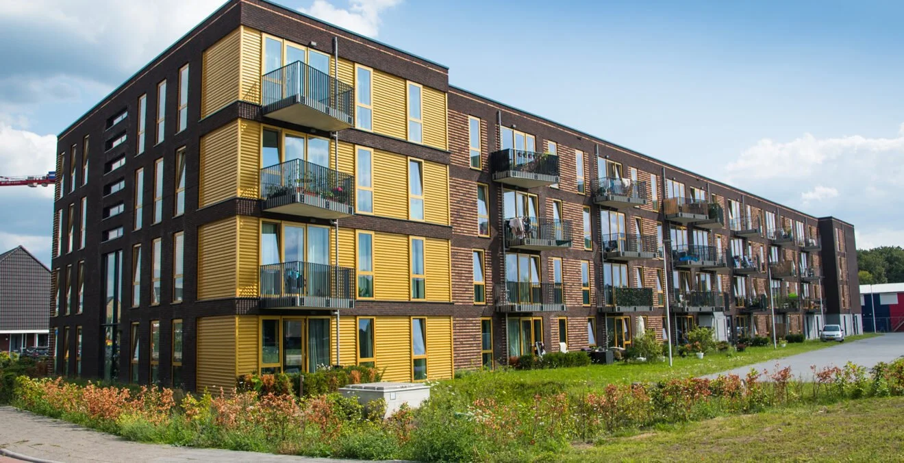 Moderne vierverdieping appartementencomplex met balkonnetjes, gevels met gele en bruine elementen, rondom groen gras en bloemen, onder een blauwe hemel met wolken.