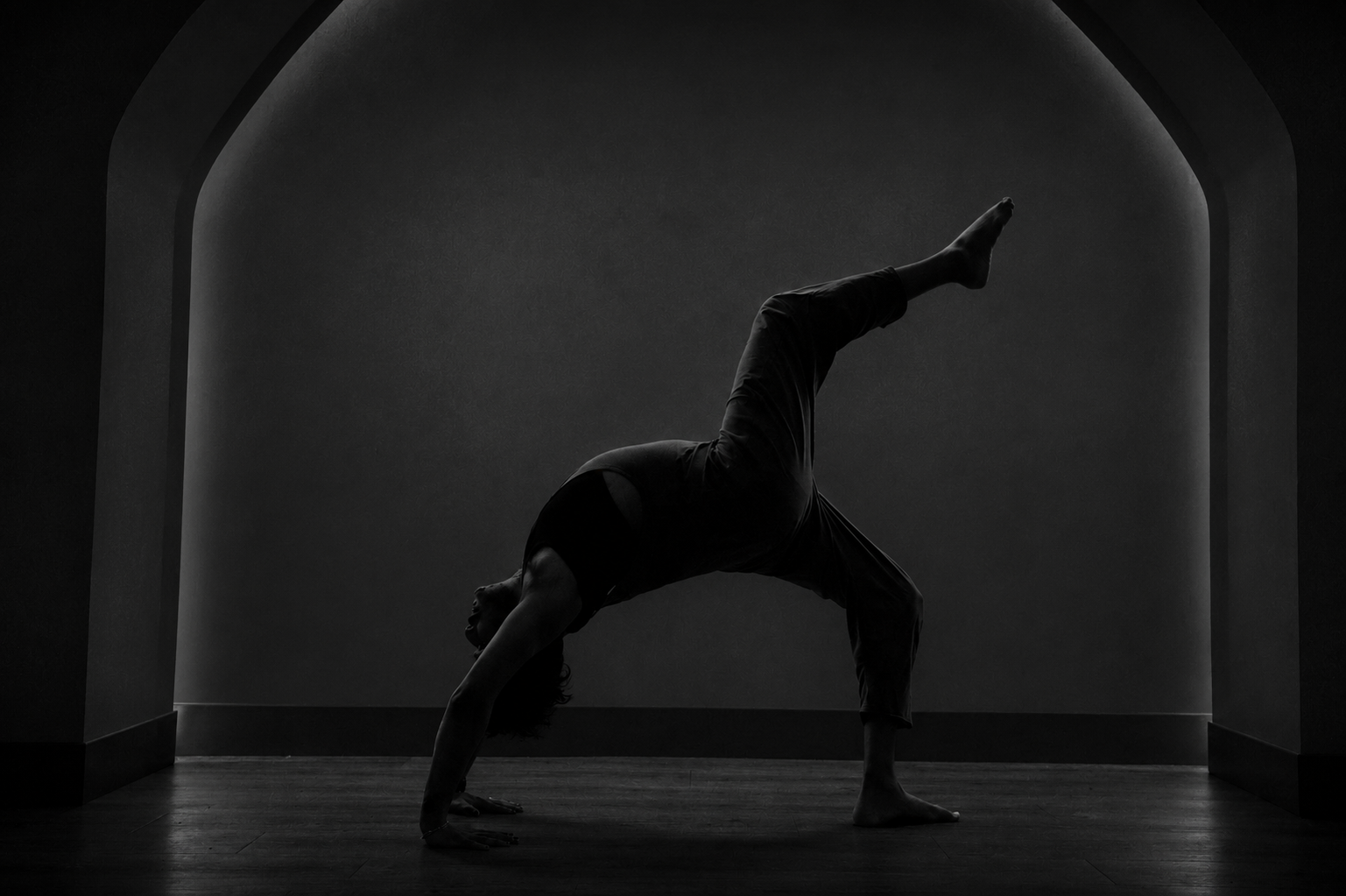 A person practicing yoga in a dimly lit room, performing a handstand with one leg extended upward. The environment features arched doorways and wooden flooring.