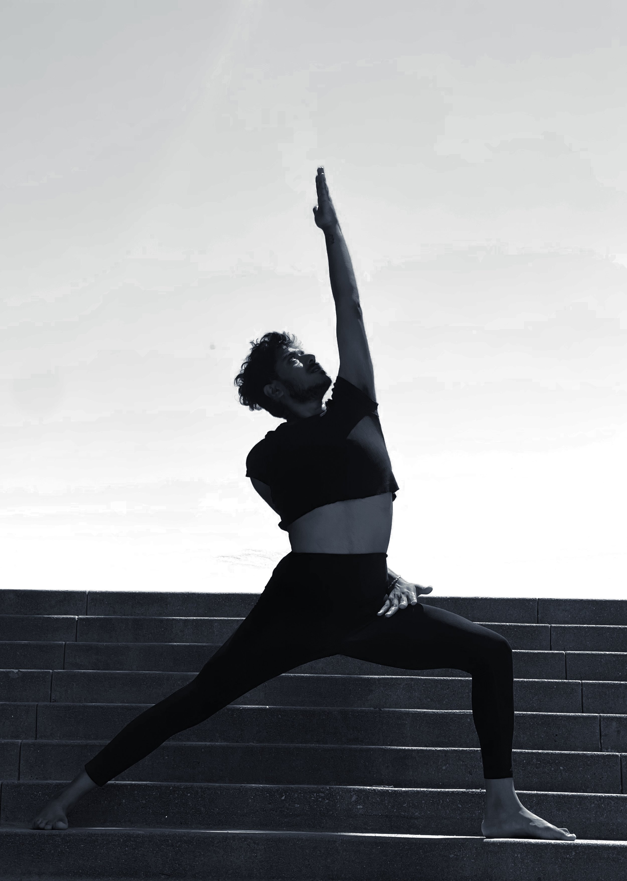 A man practicing yoga outdoors on steps, in a lunge pose with one arm reaching upwards and the other resting on his bent knee, silhouetted against a bright sky.