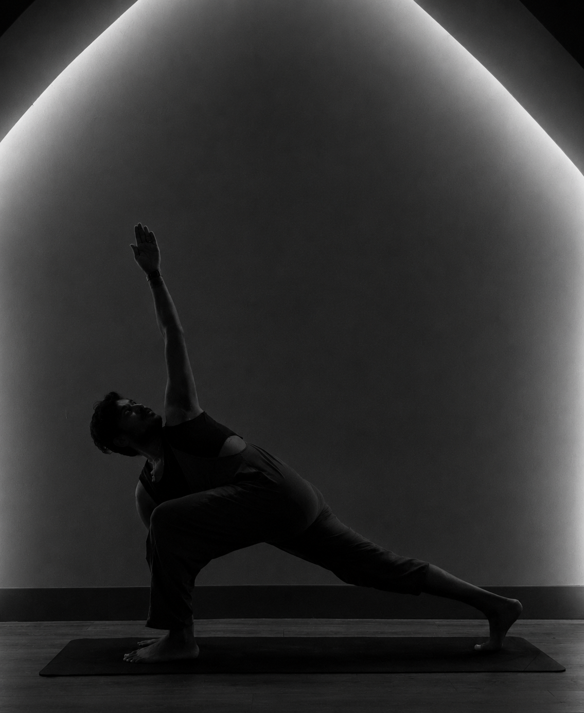 A person practicing yoga indoors, performing a side lunge pose with one arm extended upward, against a minimalist illuminated wall.