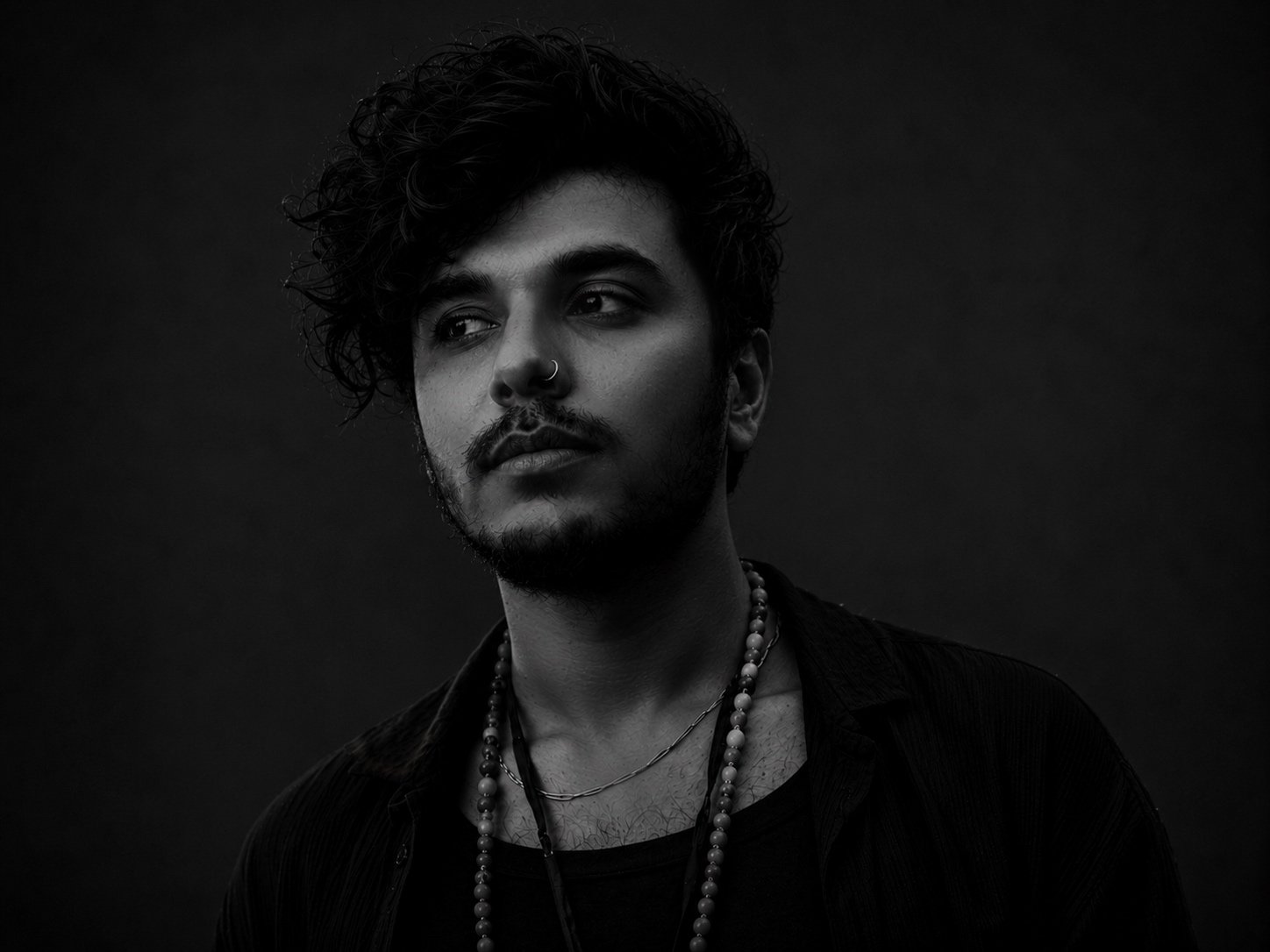 A black and white photo of a young man with curly hair wearing layered necklaces, looking to the side with a contemplative expression.