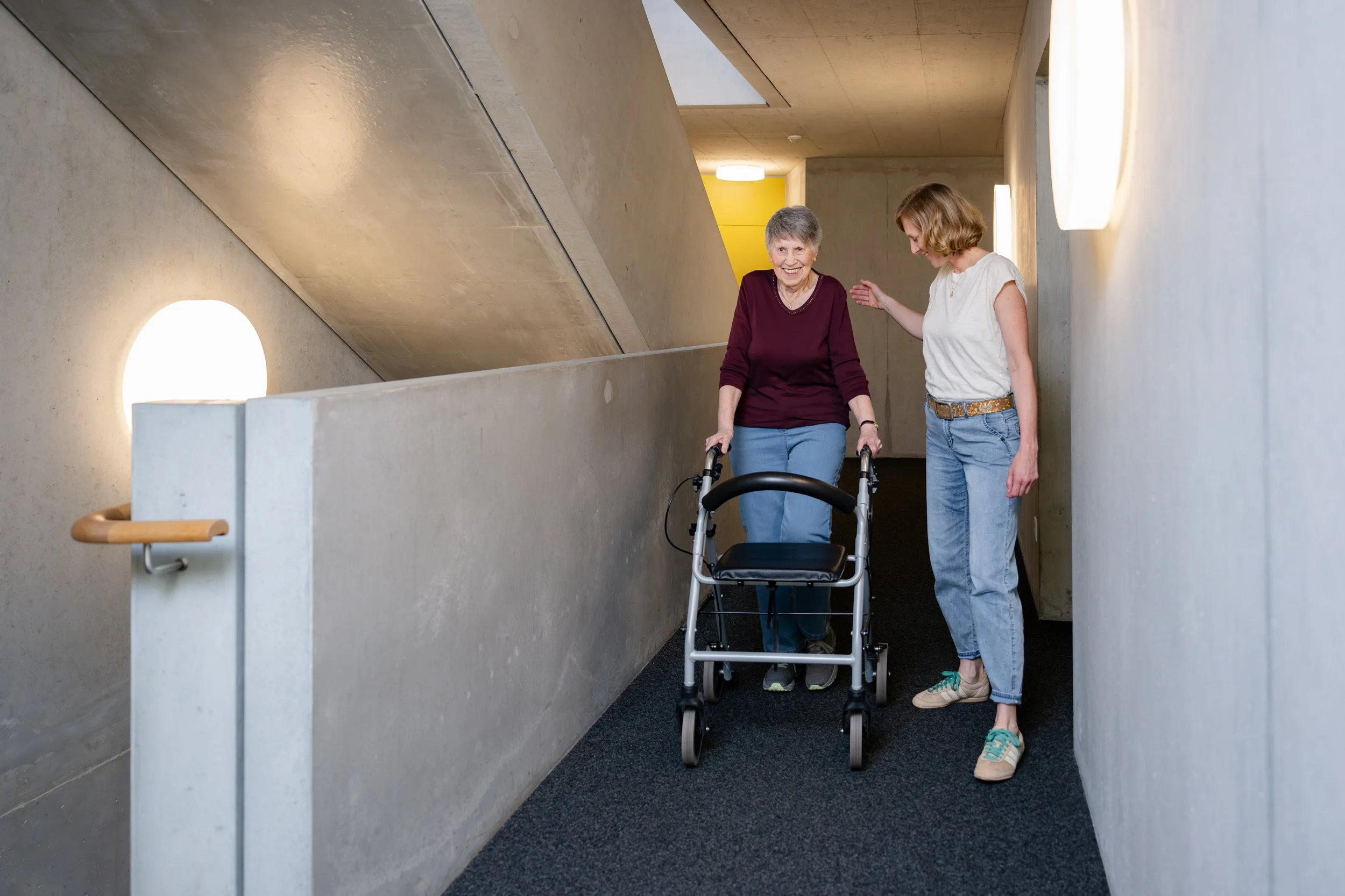 Eine ältere Frau mit Gehhilfe wird von einer jüngeren Frau in einem modernen Gebäude geführt, beide lächeln. Die Umgebung hat eine minimalistische Architektur mit Betonwänden und warmem Licht.