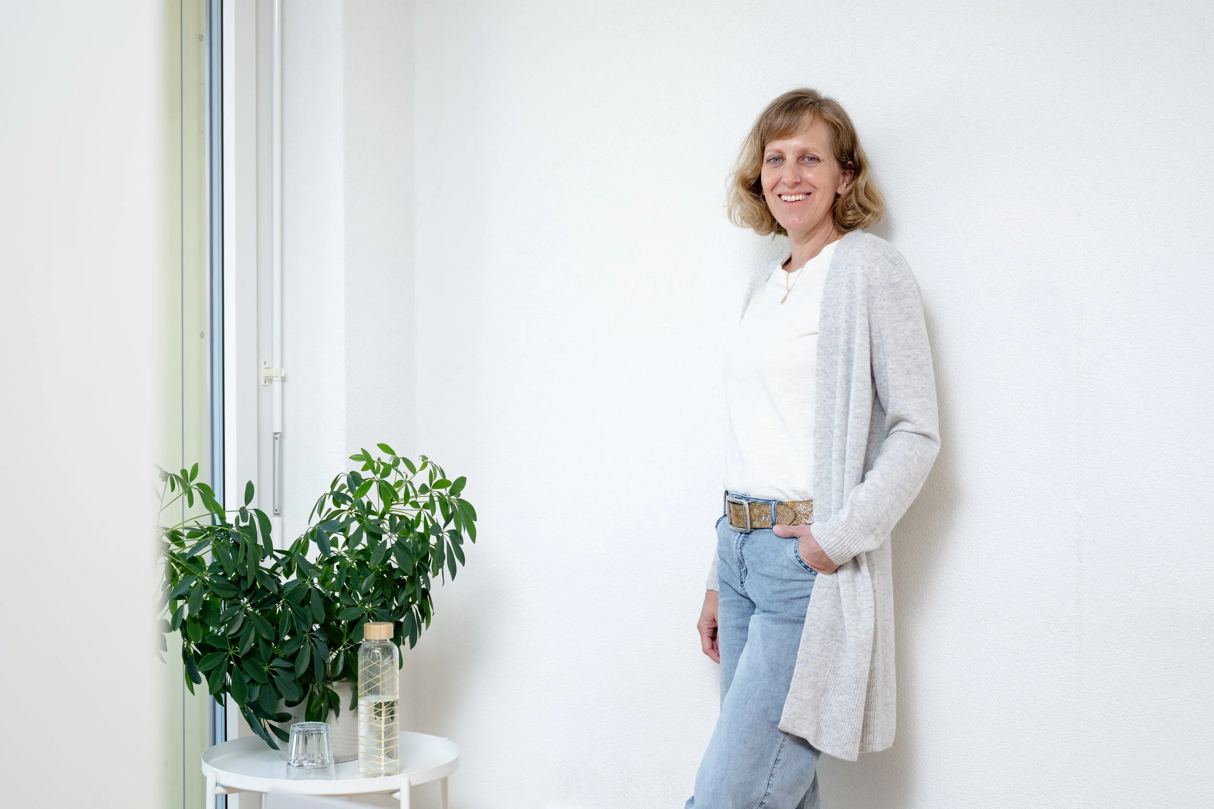 Frau steht in einem hellen Raum neben einer weißen Wand und einem Fenster, trägt eine graue Jacke, weiße Bluse und blaue Jeans, lächelt.