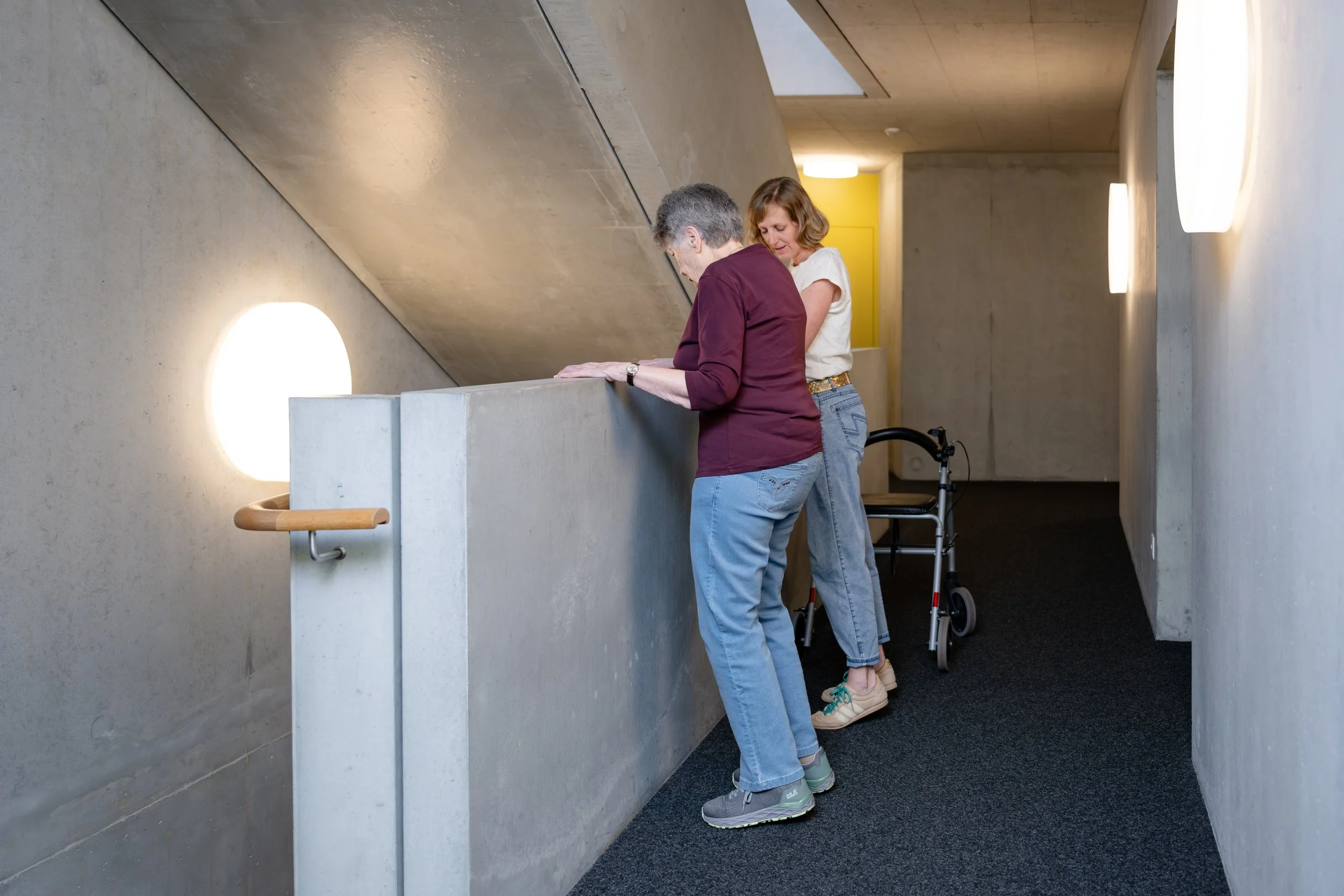 Zwei Frauen schauen über einen erhöhten Betonkiesel auf einem Flur im modernen Gebäude. Eine der Frauen benutzt einen Gehwagen, beide sind eher alte Frauen. Das Gebäude hat einen minimalistischen, modernen Stil mit hellen Wänden und Wandlampen.