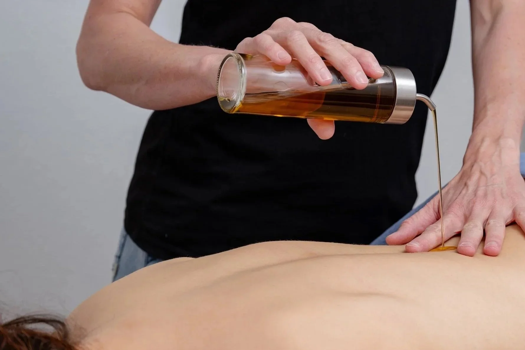 Frau liegt auf dem Bauch, während eine Person eine Flüssigkeit in einen langen, transparenten Becher gießt, vermutlich Öl, über ihrem Rücken.