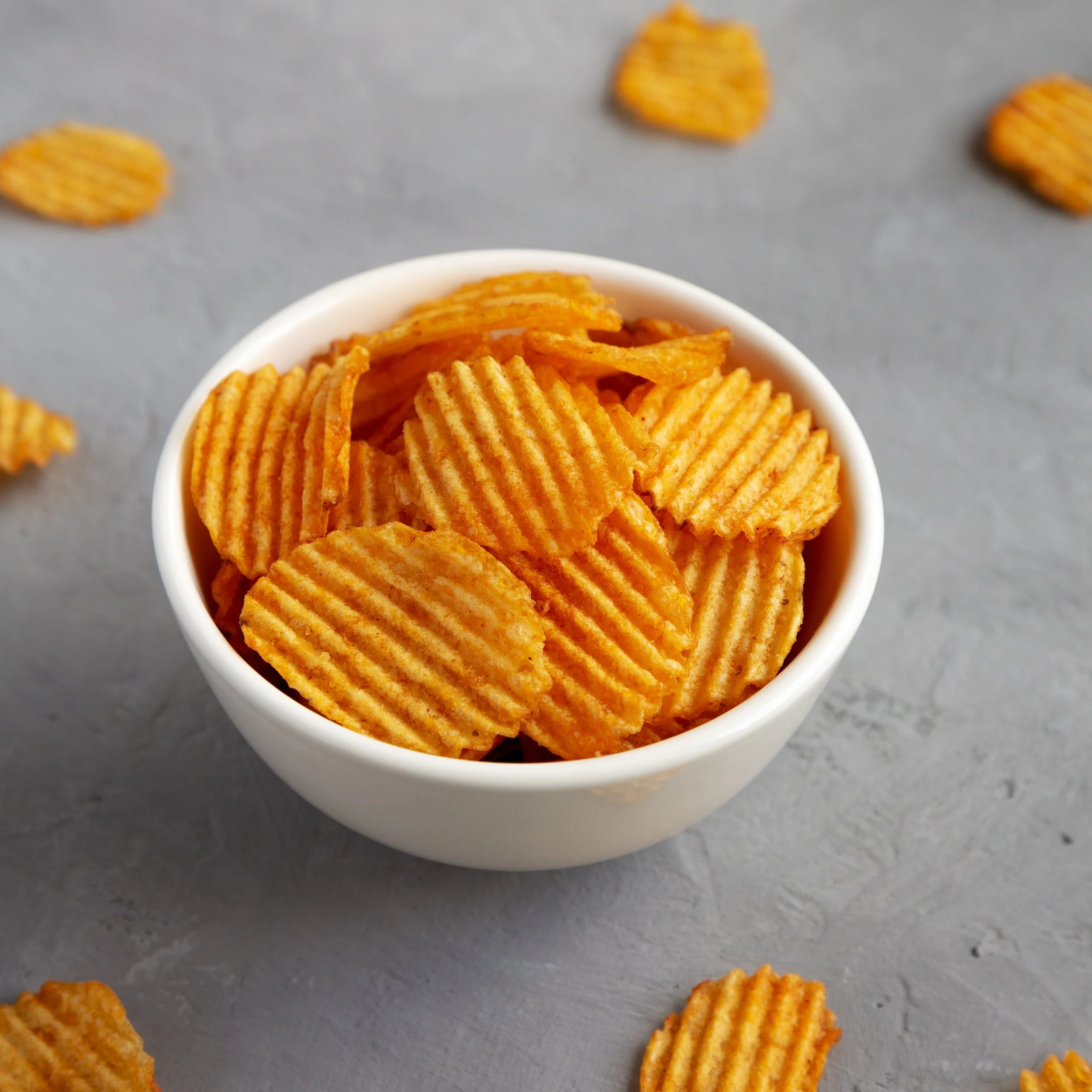 A white bowl filled with ridged, orange potato chips on a gray surface, with some chips spilled around the bowl.