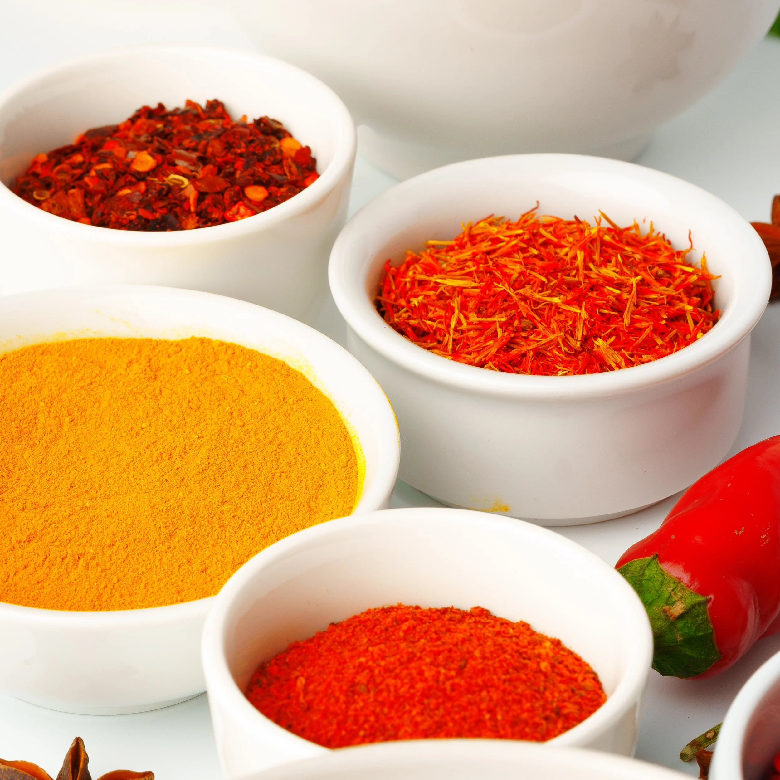 Close-up of white bowls filled with colorful spices, including red chili flakes, turmeric, and paprika, with strawberries and other ingredients partially visible.