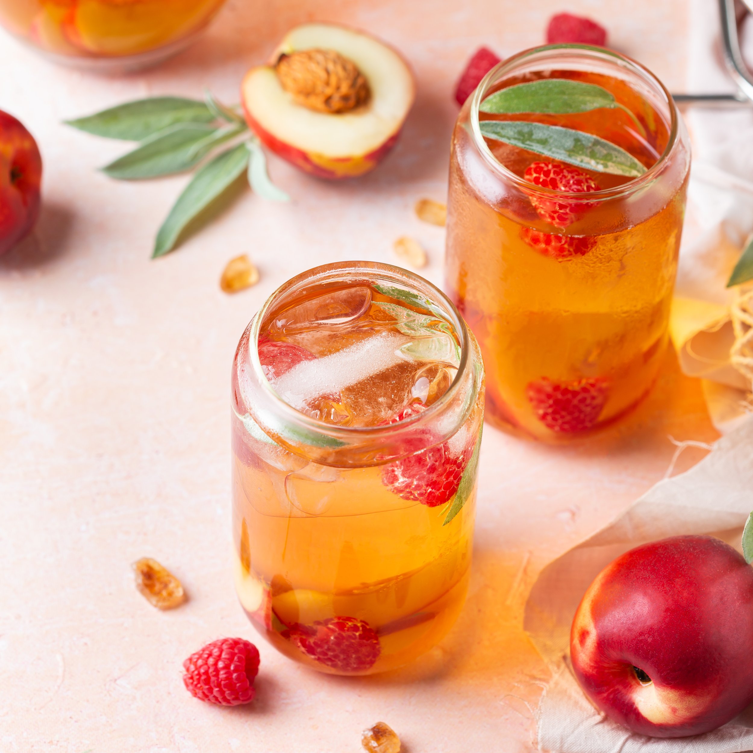 A glass jar filled with iced tea garnished with strawberries and mint leaves, with sliced peaches, raspberries, peaches, and some green leaves scattered around on a light-colored surface.
