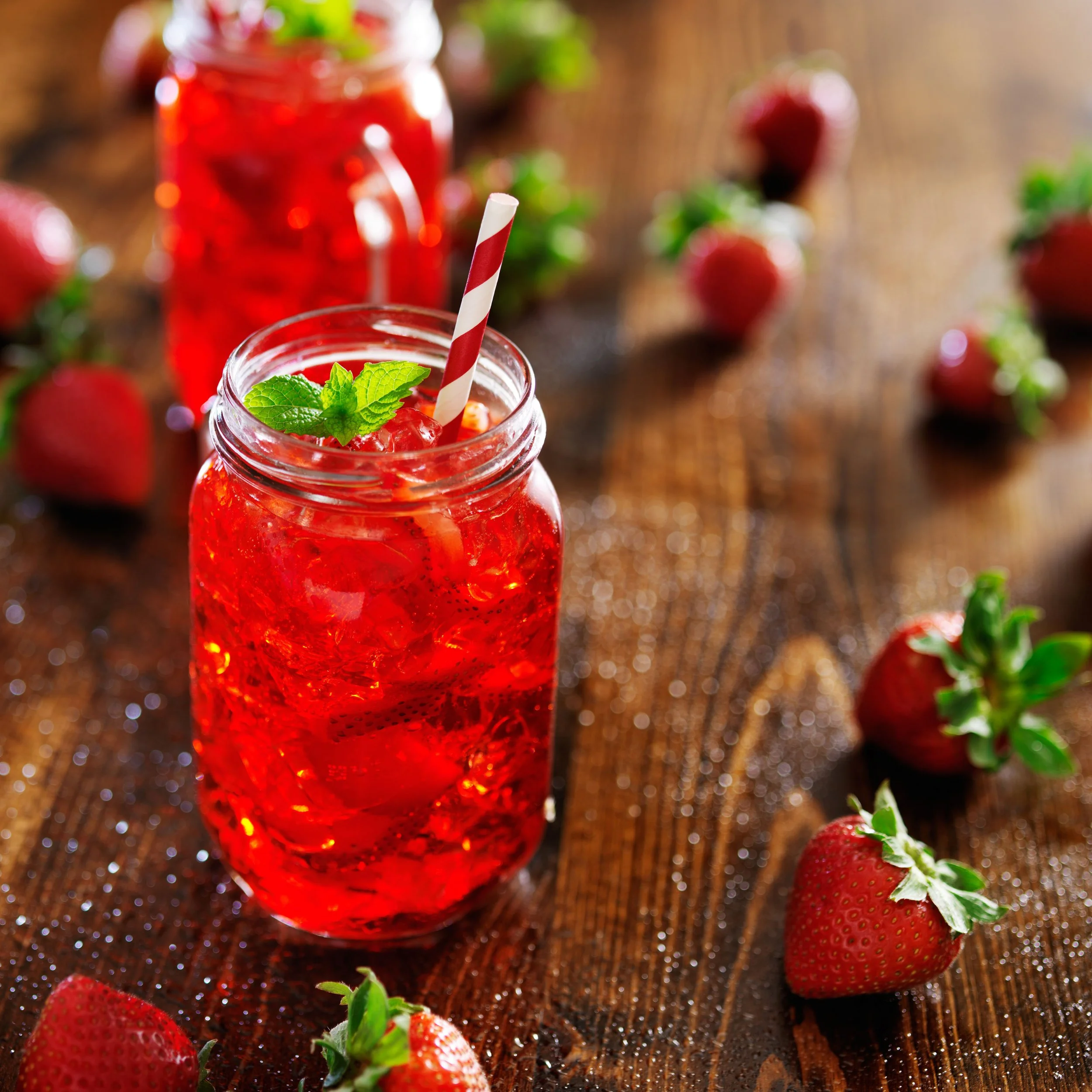 Close-up of strawberries with a jar of strawberry-infused drink, garnished with mint leaves and a striped straw, on a wooden surface.