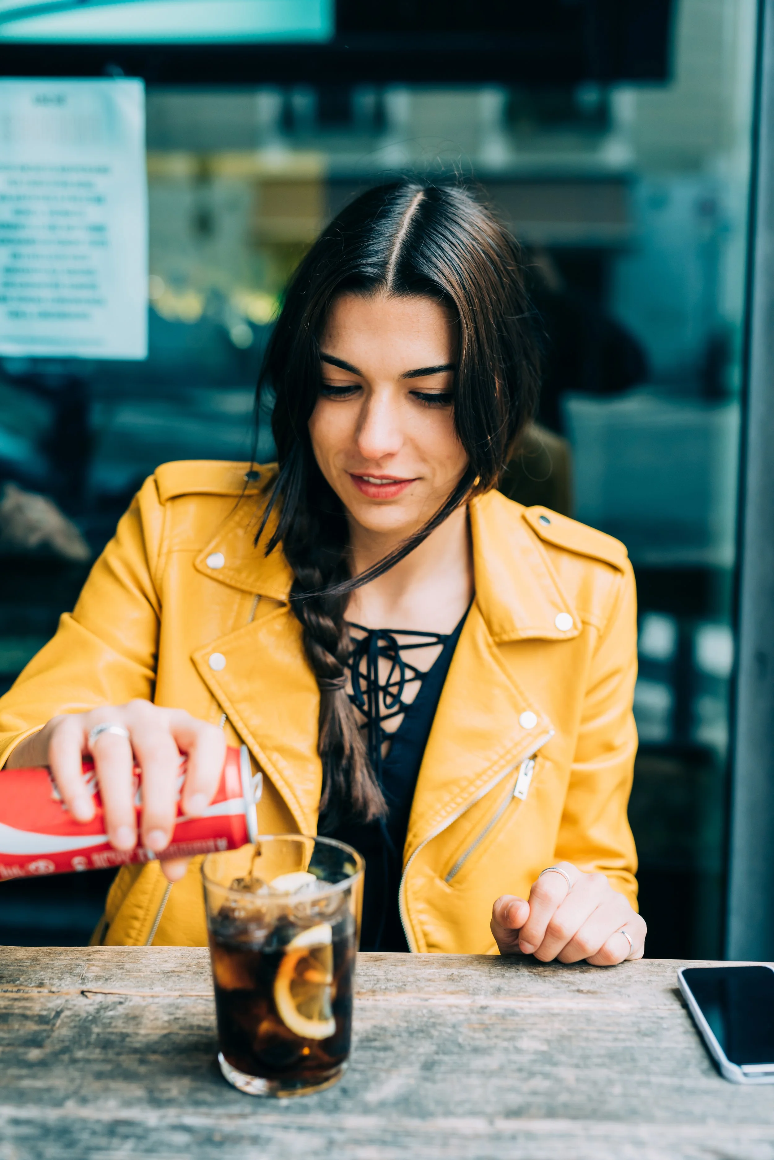 A young woman with dark hair wearing a yellow jacket is pouring a drink with lemon slices into a glass of soda, sitting at a rustic wooden table.