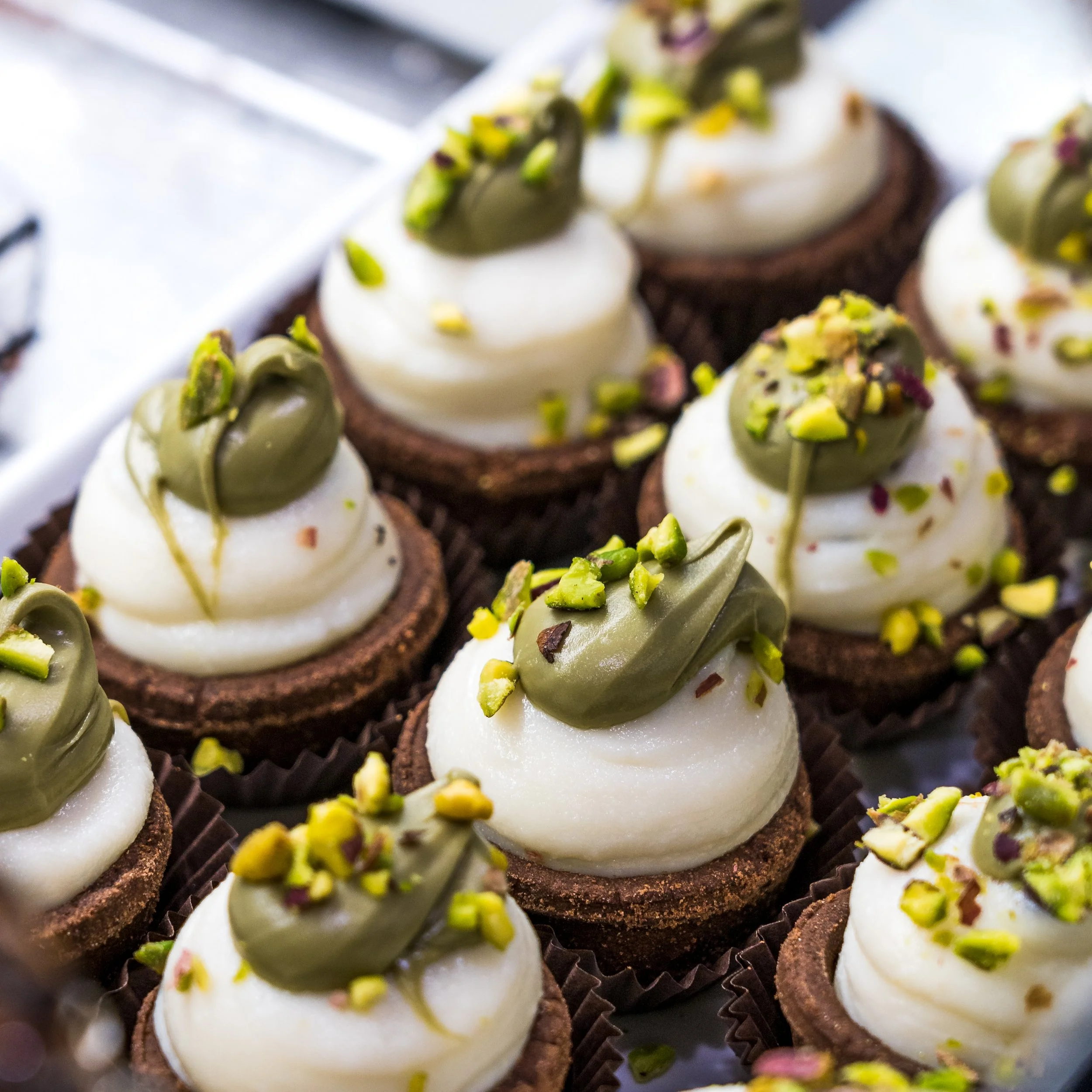 Close-up of multiple mini chocolate tart desserts topped with white cream, green pistachio pieces, and green drizzle.
