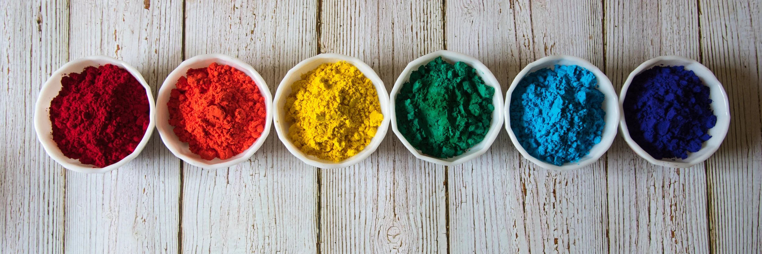 Six white bowls containing powdered pigments in red, orange, yellow, green, blue, and dark blue arranged in a row on a wooden surface.