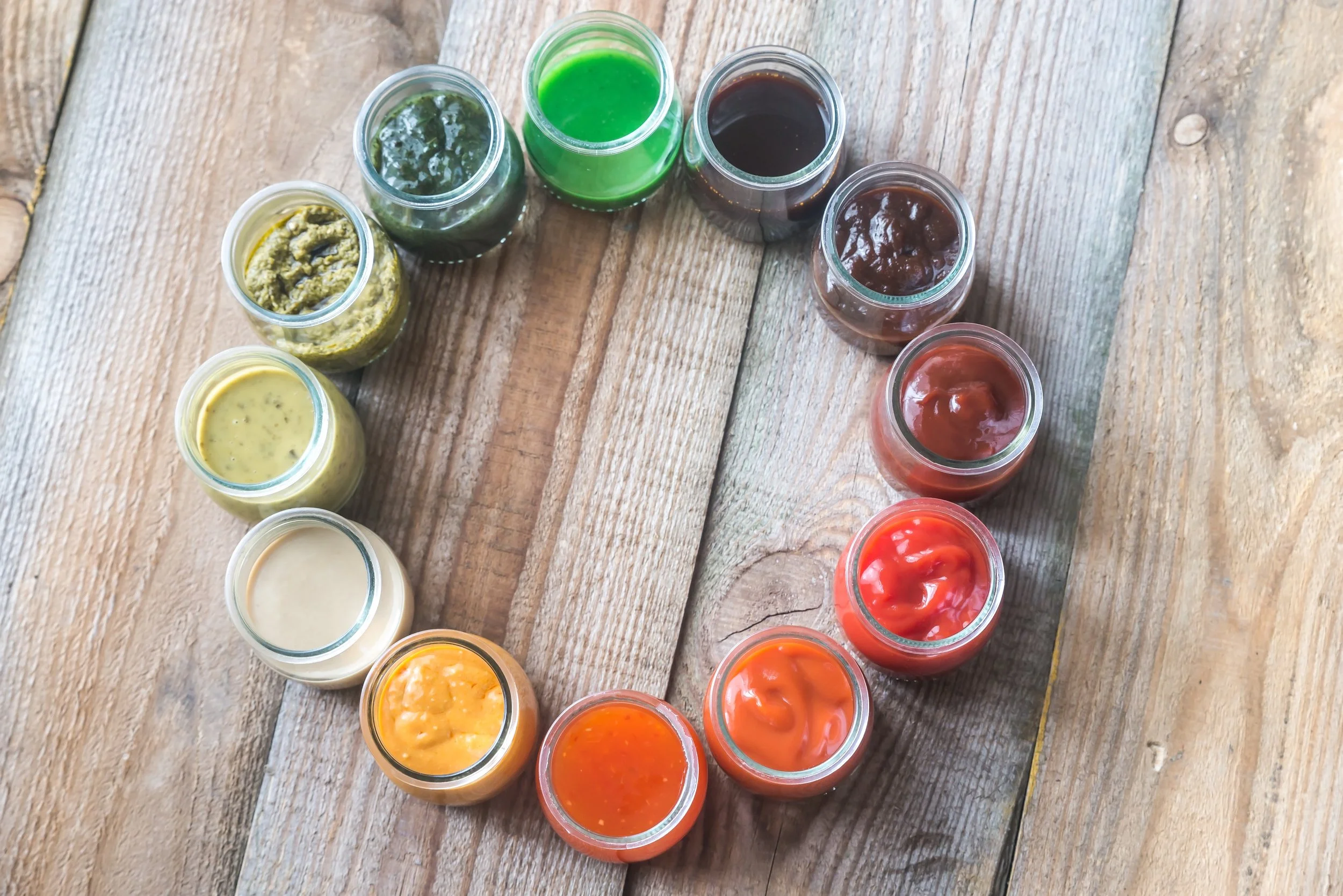 A top view of multiple small glass jars containing various colorful sauces or condiments arranged on a wooden surface.
