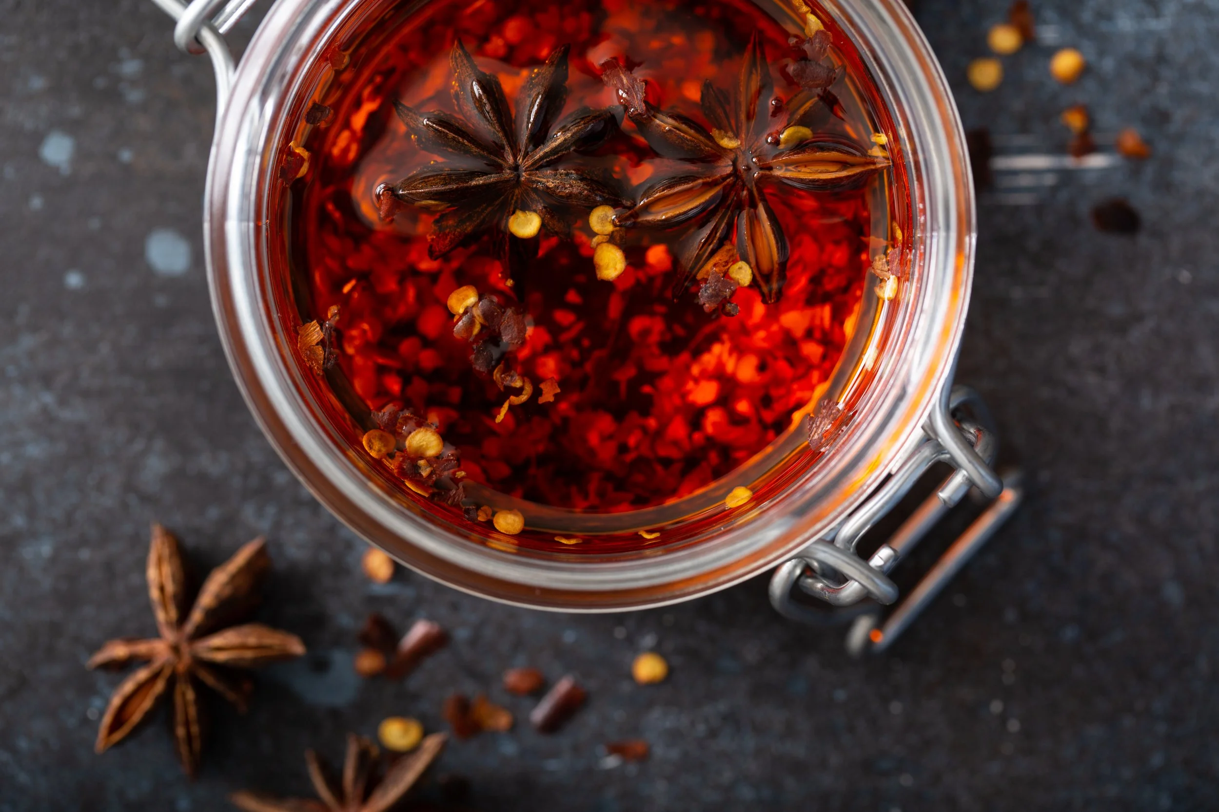 A glass jar filled with a dark red liquid containing star anise, red chili flakes, and peppercorns, placed on a dark surface with star anise and peppercorns scattered around.