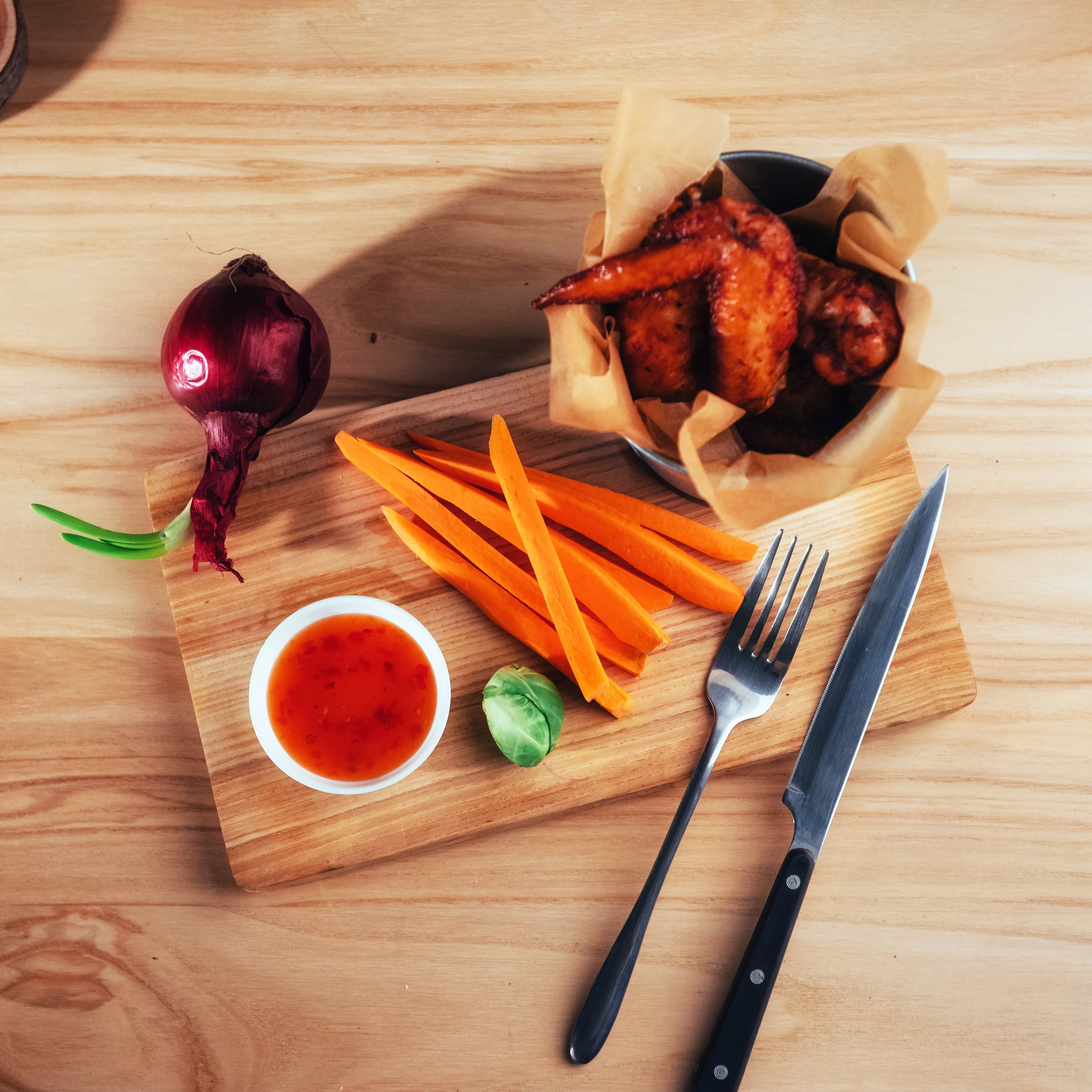 A wooden cutting board with a red onion, a small bowl of orange dipping sauce, and brightly colored carrot sticks. There is a knife and fork on the right side of the image, and a small piece of green vegetable, possibly a basil leaf, near the bowl.
