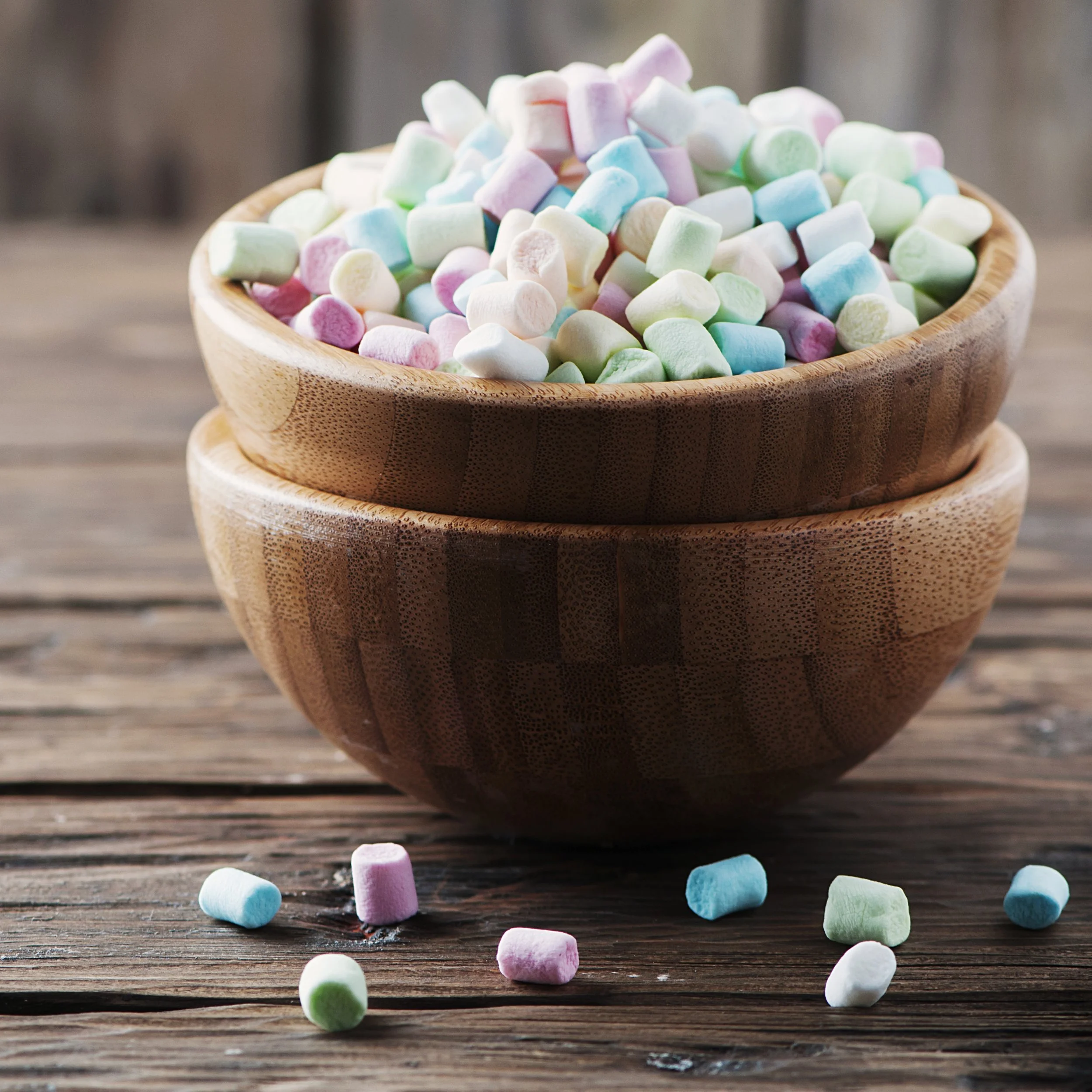Colorful mini marshmallows in shades of pink, green, blue, and white in a wooden bowl on a rustic wooden surface with some marshmallows scattered around.