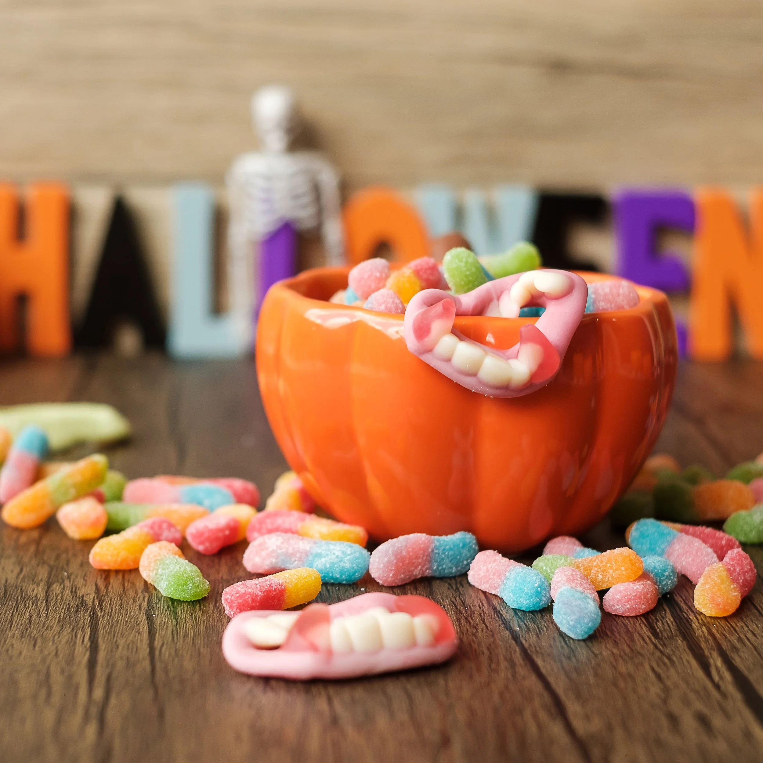 Colorful sour gummy candies in a small orange bowl, with some candies scattered on a wooden surface. In the background, there are blurred colorful letters and a blurred silver skeleton figure.