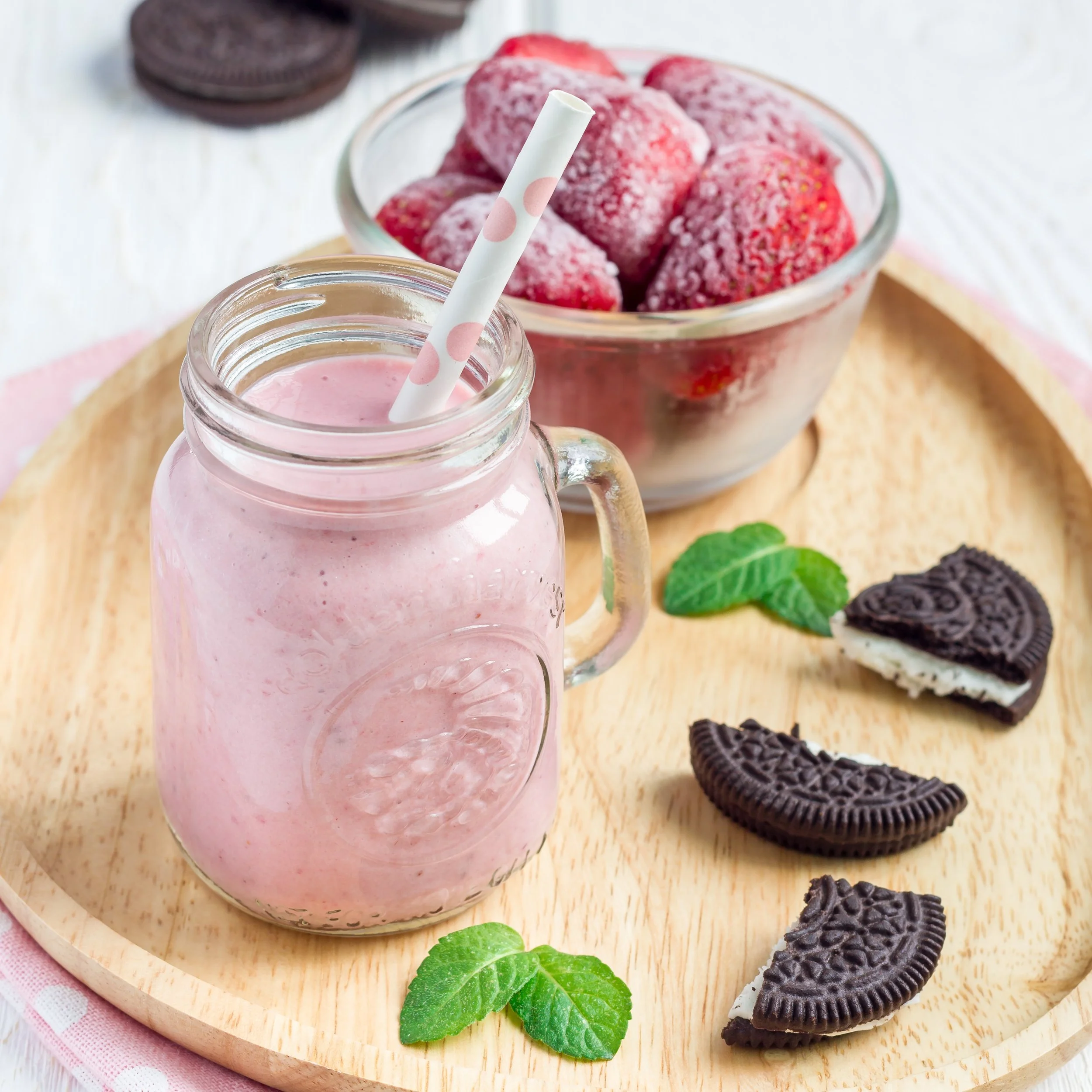 A glass bottle of pink strawberry smoothie with a white and pink polka dot straw, a bowl of frozen strawberries, and Oreo cookies arranged on a wooden tray with green mint leaves.