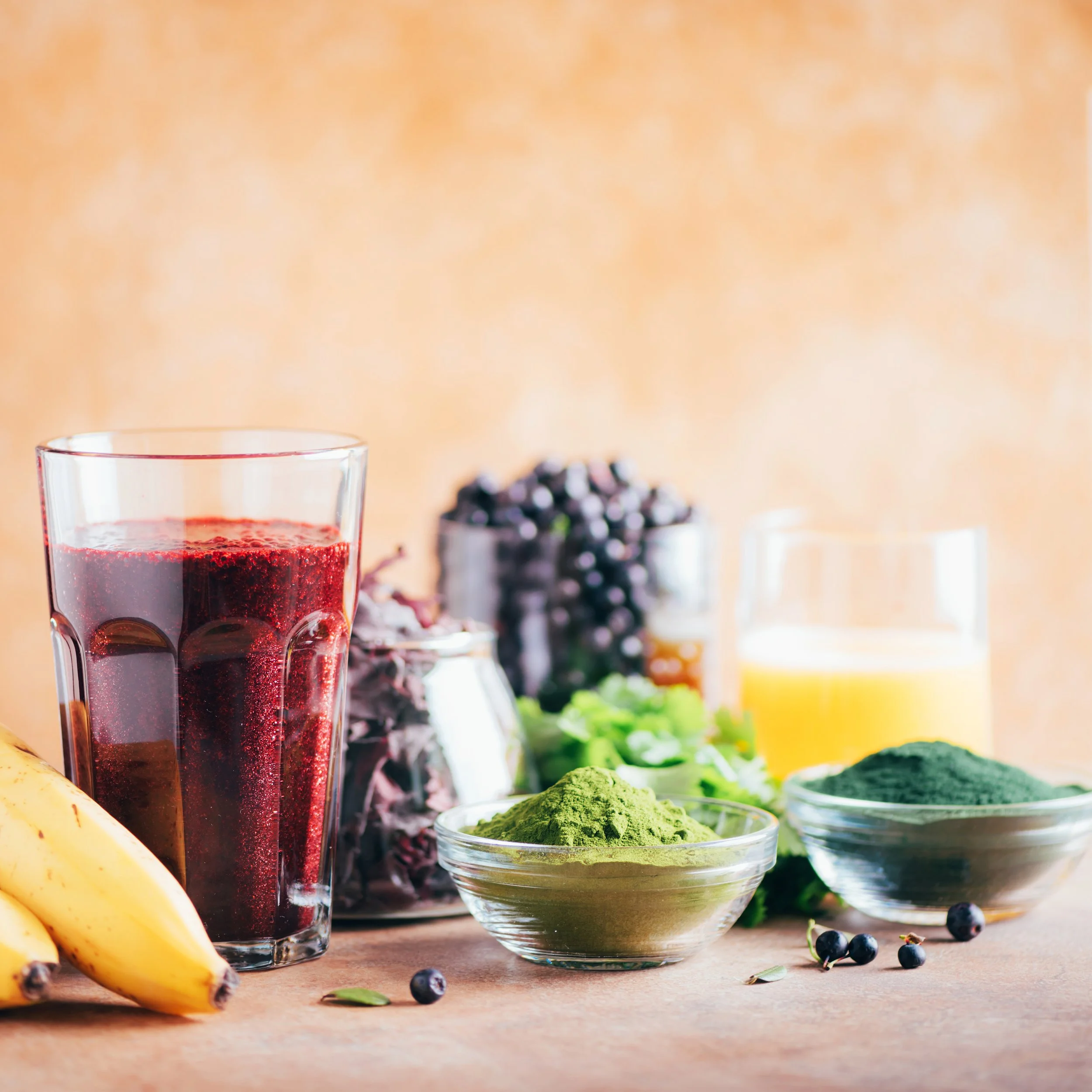 A glass of dark red berry juice, a bowl of green powder, bananas, blueberries, and various fresh berries and greens on a wooden surface with a blurred background.