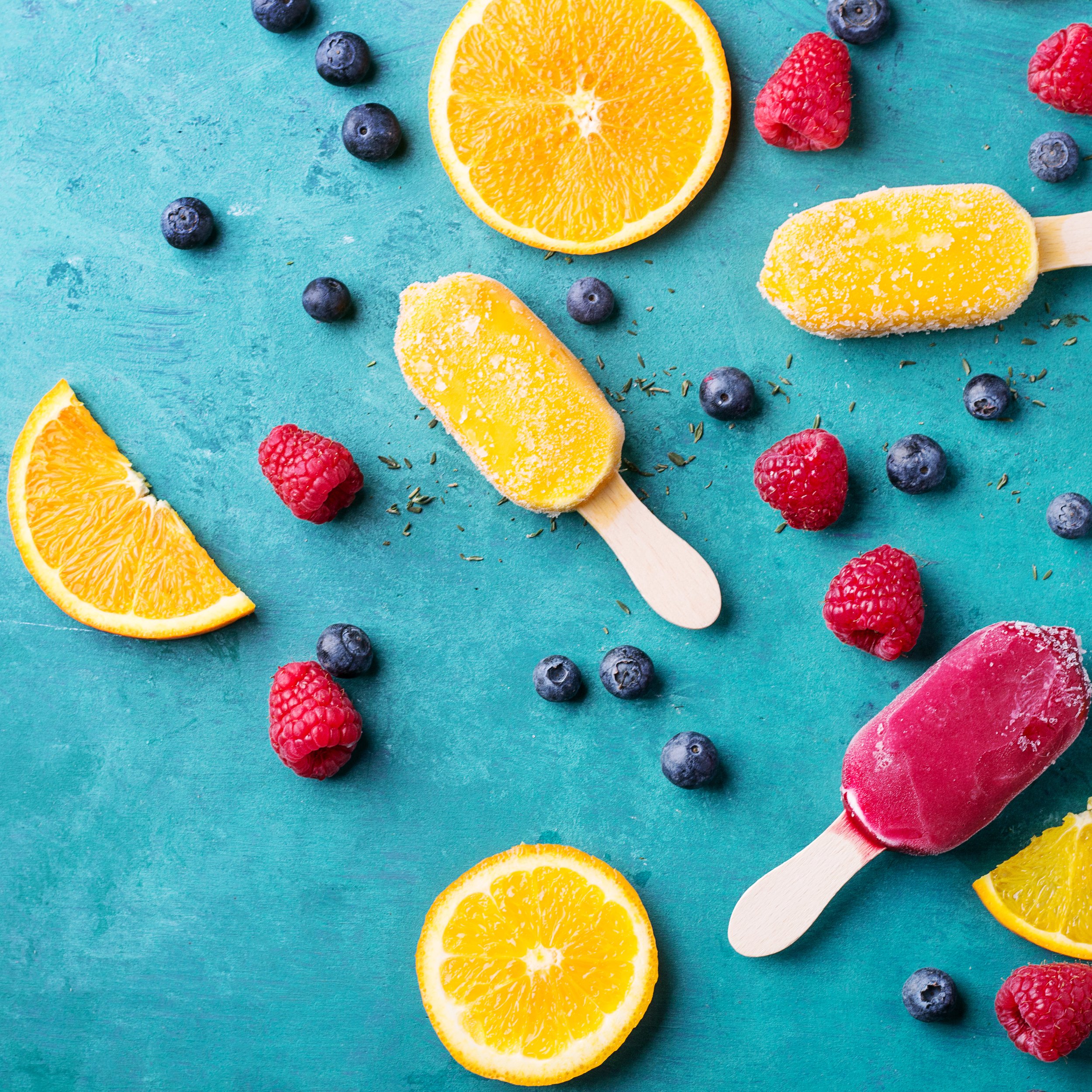 Slices of orange and lemon, raspberries, blueberries, and ice pops on a teal background.