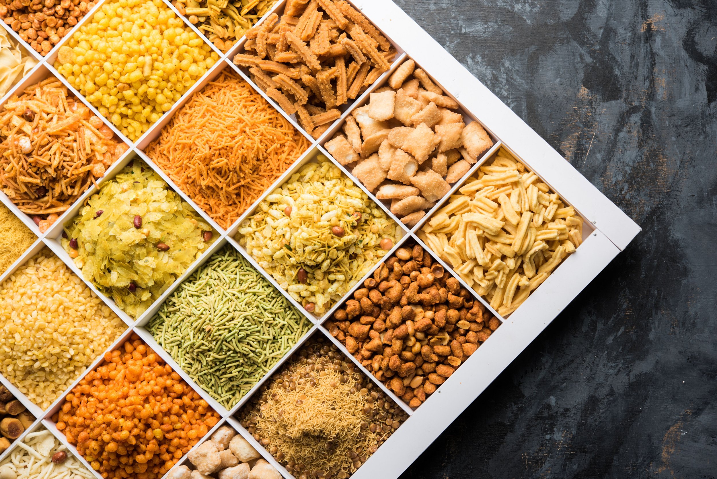 A white box with multiple compartments filled with various colorful Indian snacks and savory treats, placed on a dark textured surface.