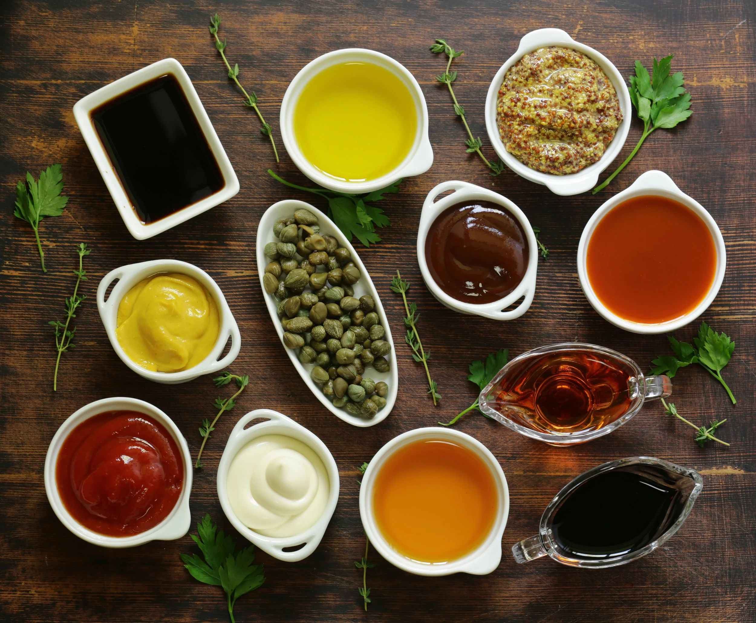 Assorted sauces and condiments in small white bowls on a wooden table, decorated with sprigs of fresh herbs.