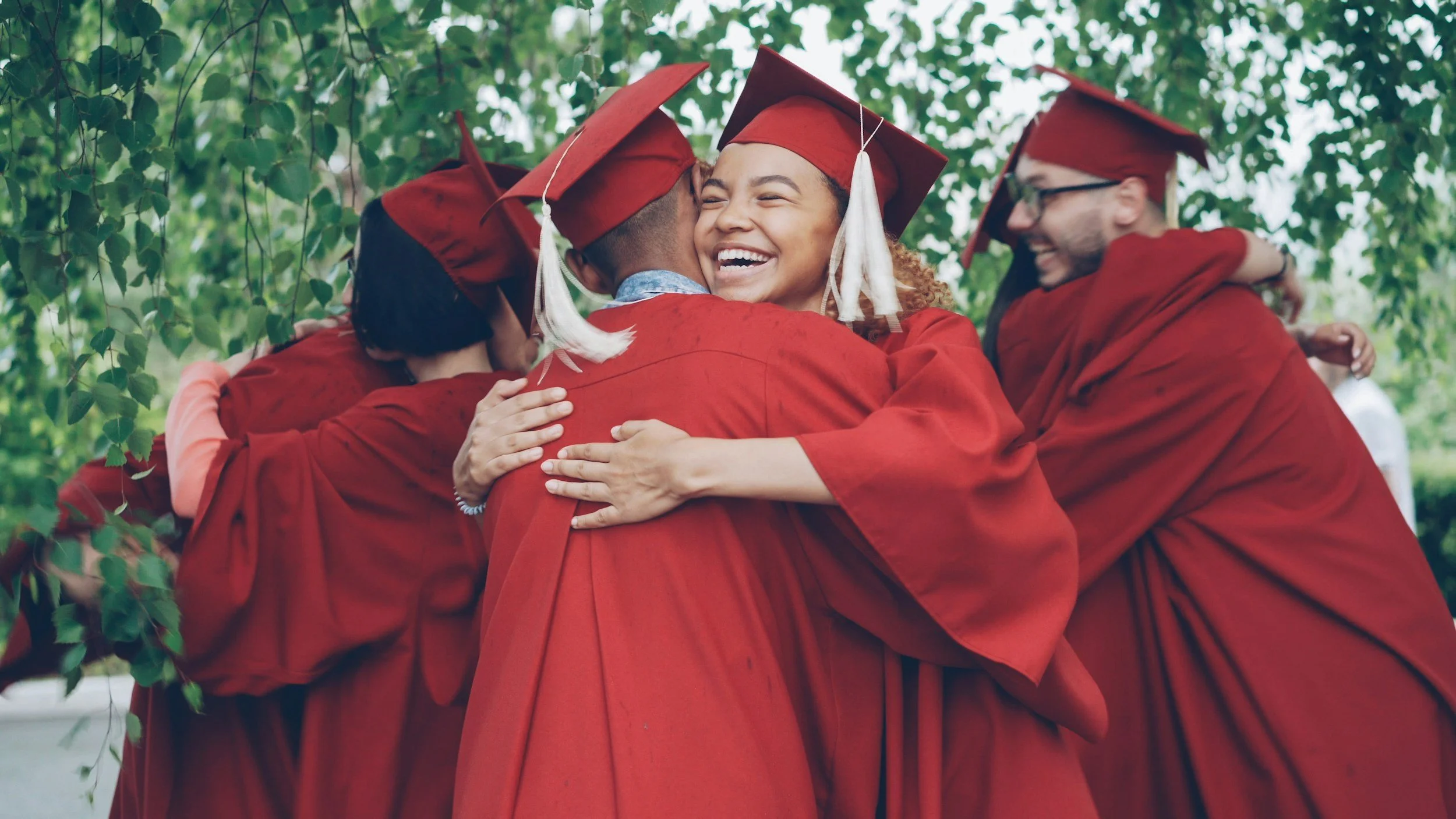 Group of graduates in red caps and gowns hugging outdoors.