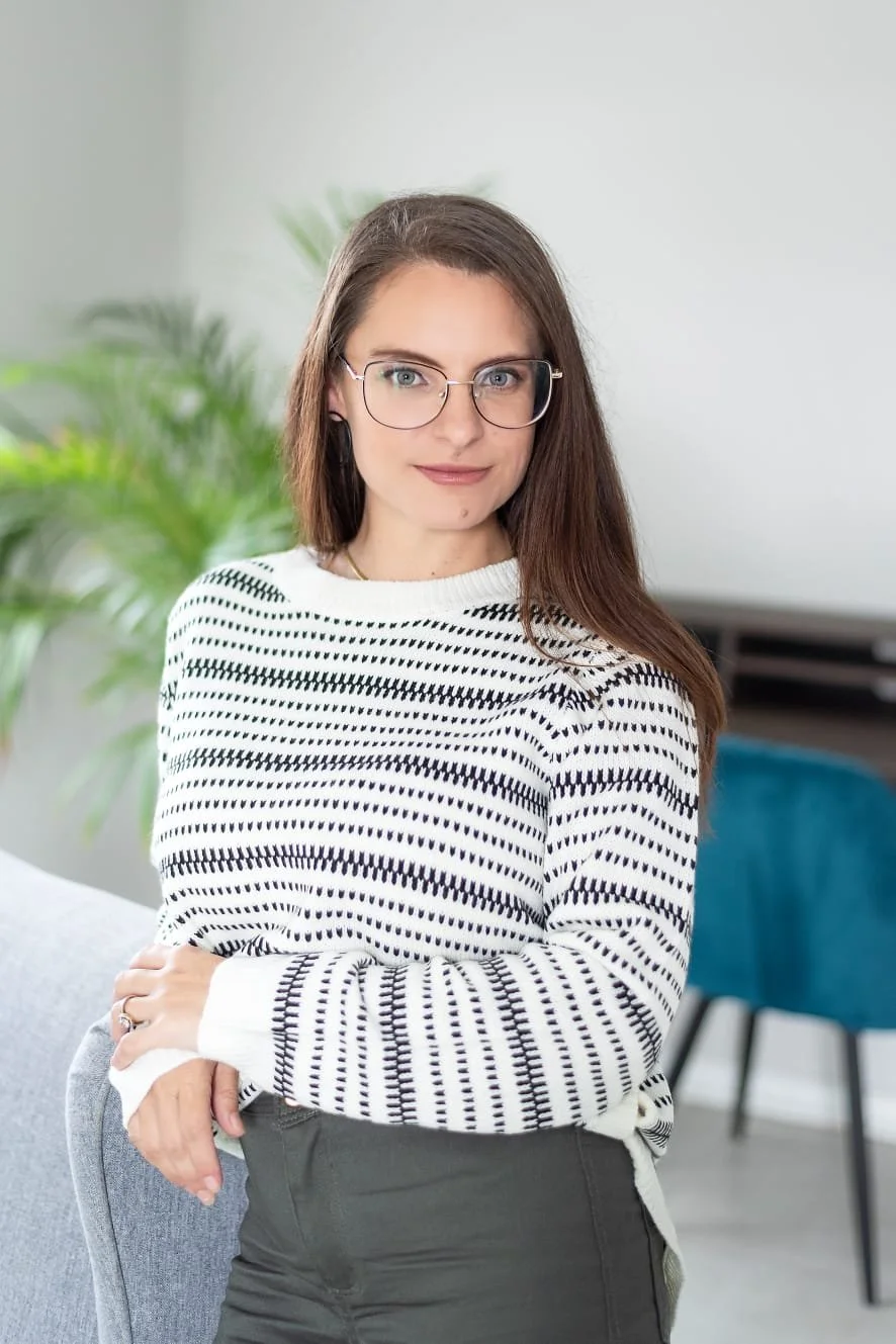 A young woman with brown hair and glasses, wearing a white sweater with black horizontal stripes, standing indoors near a green plant and a light-colored wall.