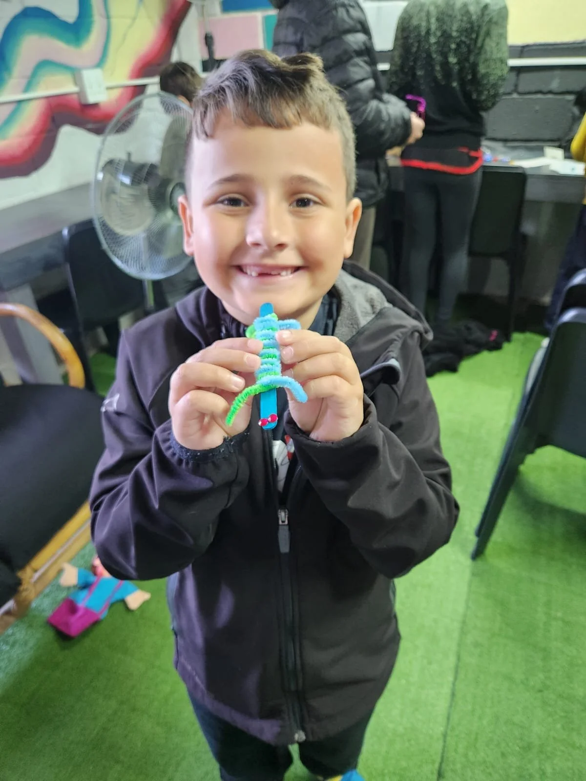 A young boy smiling and holding a colorful pipe cleaner craft shaped like a sword, standing on a green carpet in a room with other people in the background.
