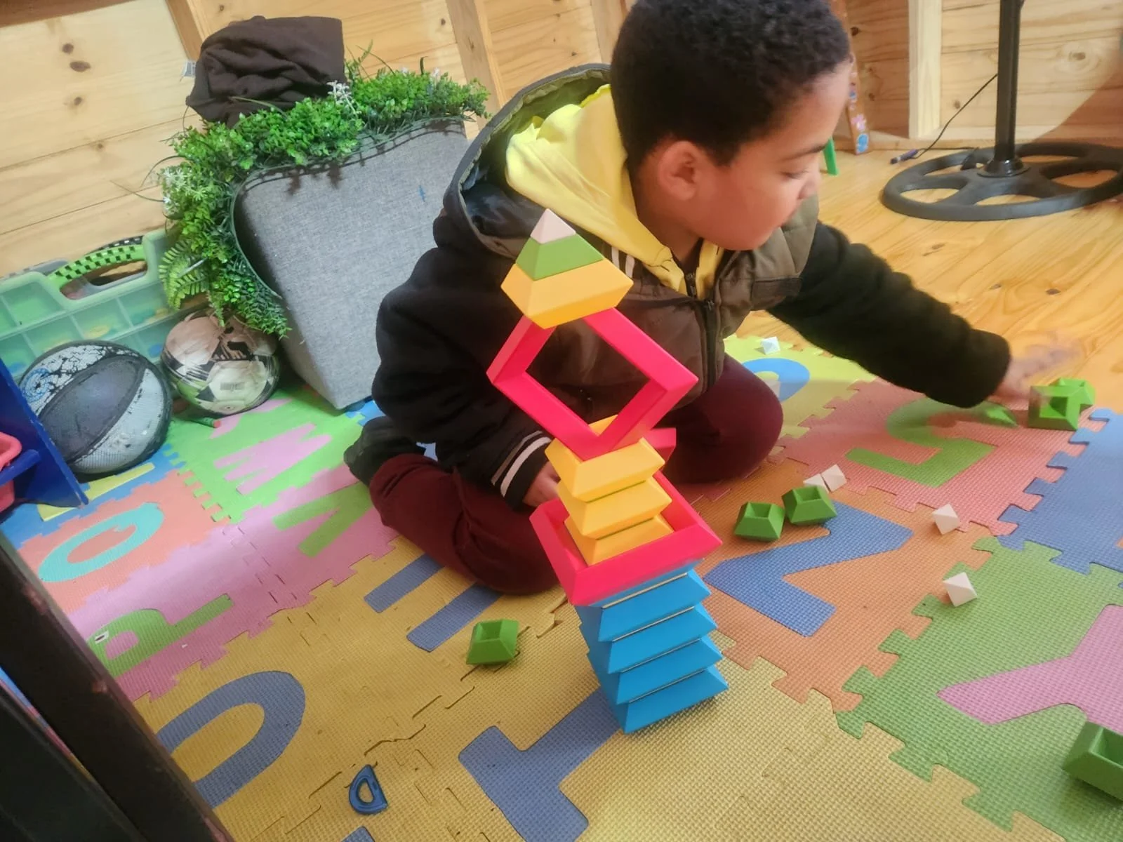 A young boy with dark curly hair playing with colorful geometric stacking blocks on a foam play mat. The room has wooden walls and a cozy atmosphere.