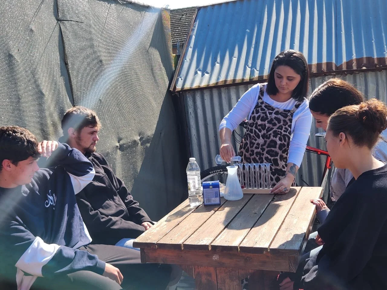 Four young people sitting at a wooden table outdoors, with a woman demonstrating a science experiment with beakers and a clear liquid, while the three others observe. The setting appears to be a backyard with metal and tarp structures.