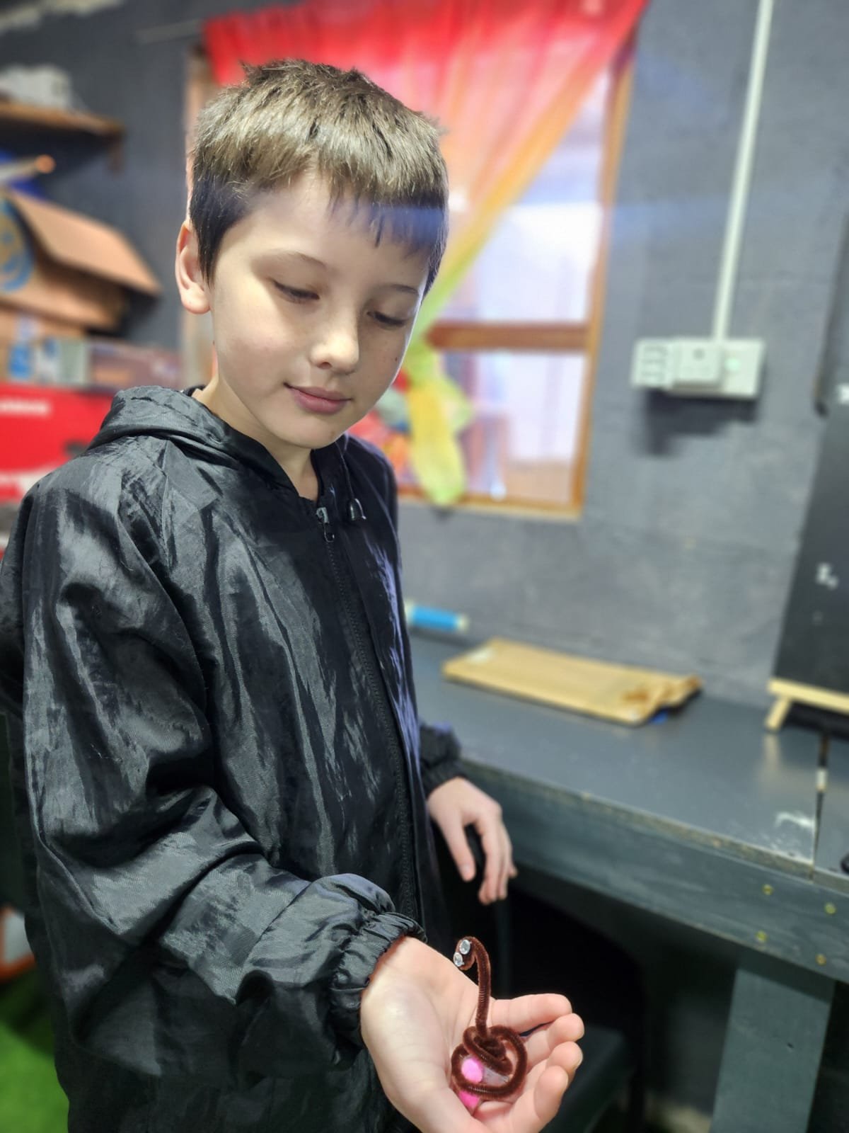 A boy with short brown hair and wearing a black jacket is holding a small, brown, curly object in his hand. He is looking down at it with a gentle expression. The background contains a colorful curtain, shelves with boxes, and a wooden plank on a table inside a room with gray walls.