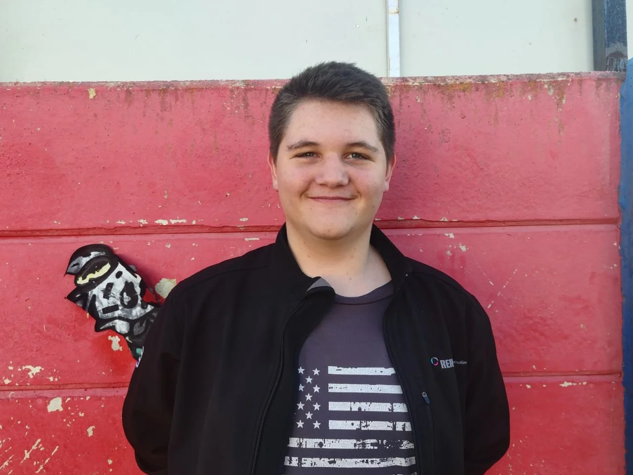 Young man with short dark hair smiling, wearing a black jacket and a t-shirt with a distressed American flag design, standing in front of a red wall with some peeling paint and graffiti.